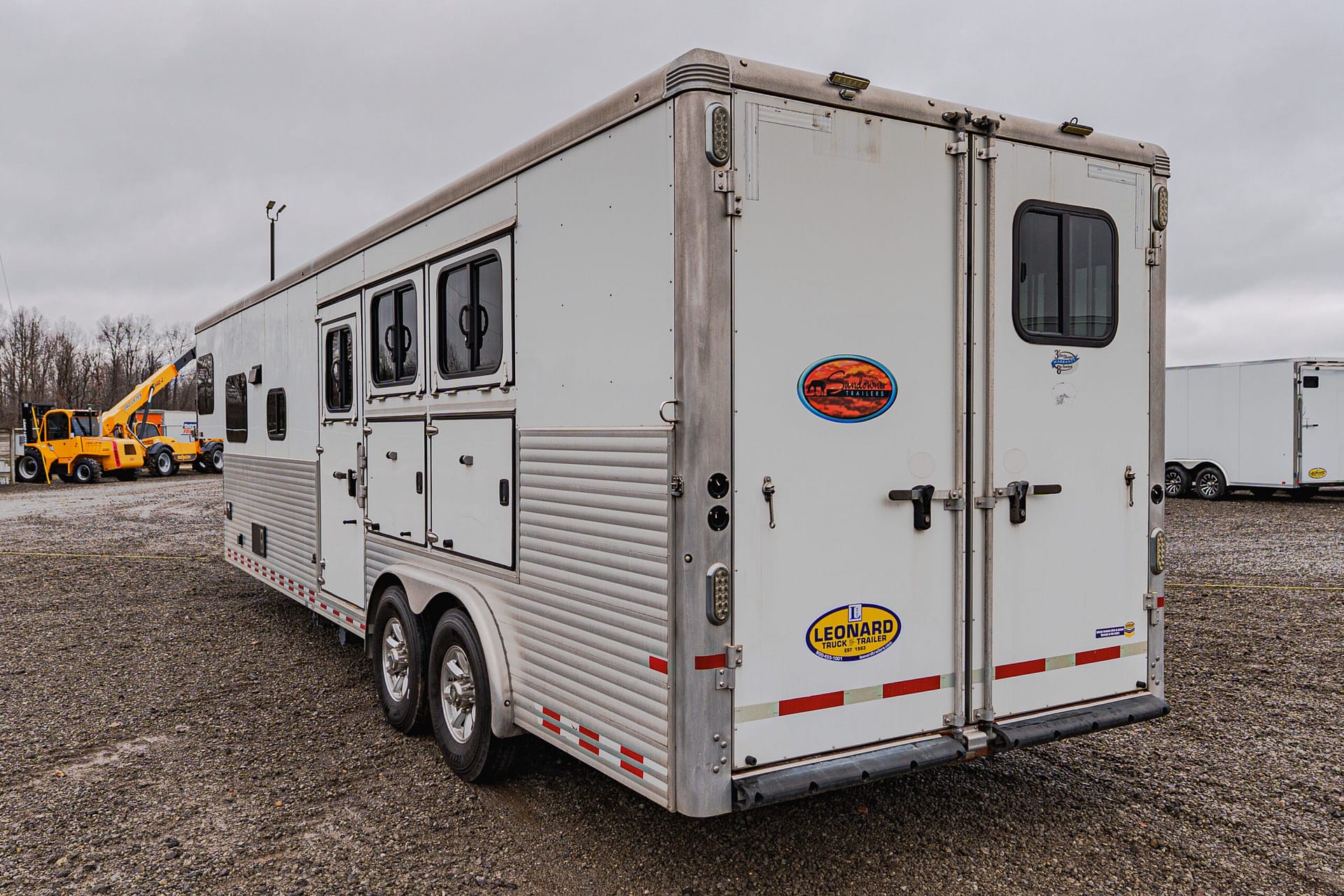 Rear three-quarter view of a large silver trailer with windows and decals, parked on a gravel lot with yellow construction equipment in the background.