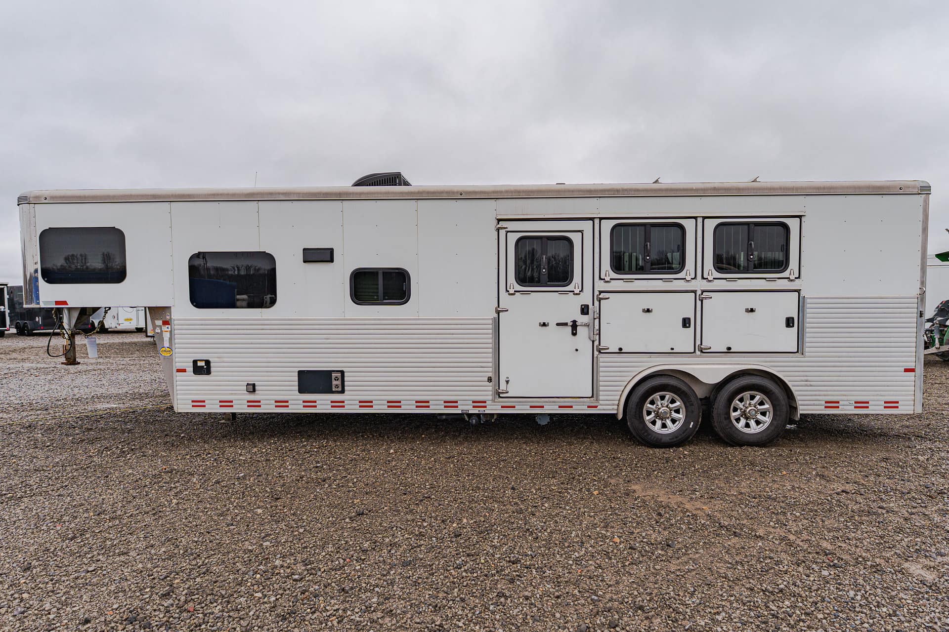 White travel trailer with multiple windows and two axles parked on a gravel lot under a cloudy sky