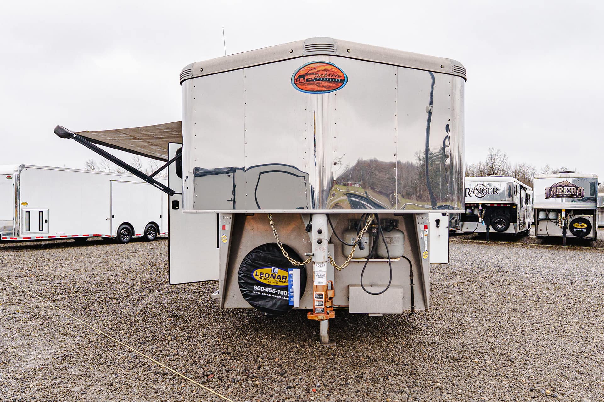 Rear view of a polished aluminum semi-trailer in a truck yard, with other trailers in the background and a spare tire covered by a Leonard brand protector in the foreground