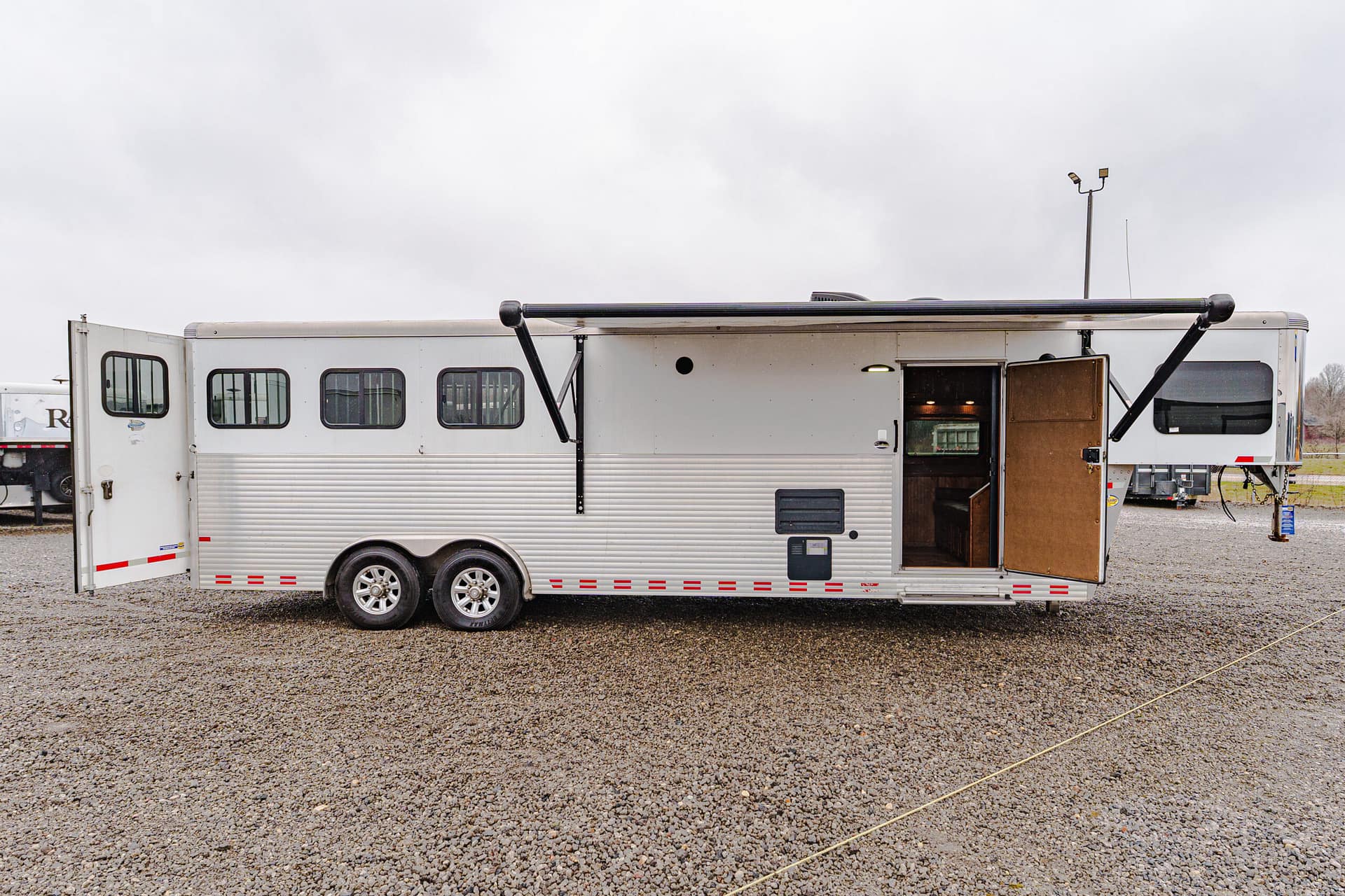 White travel trailer with open door and extended awning on a gravel lot; other trailers visible in background under cloudy sky