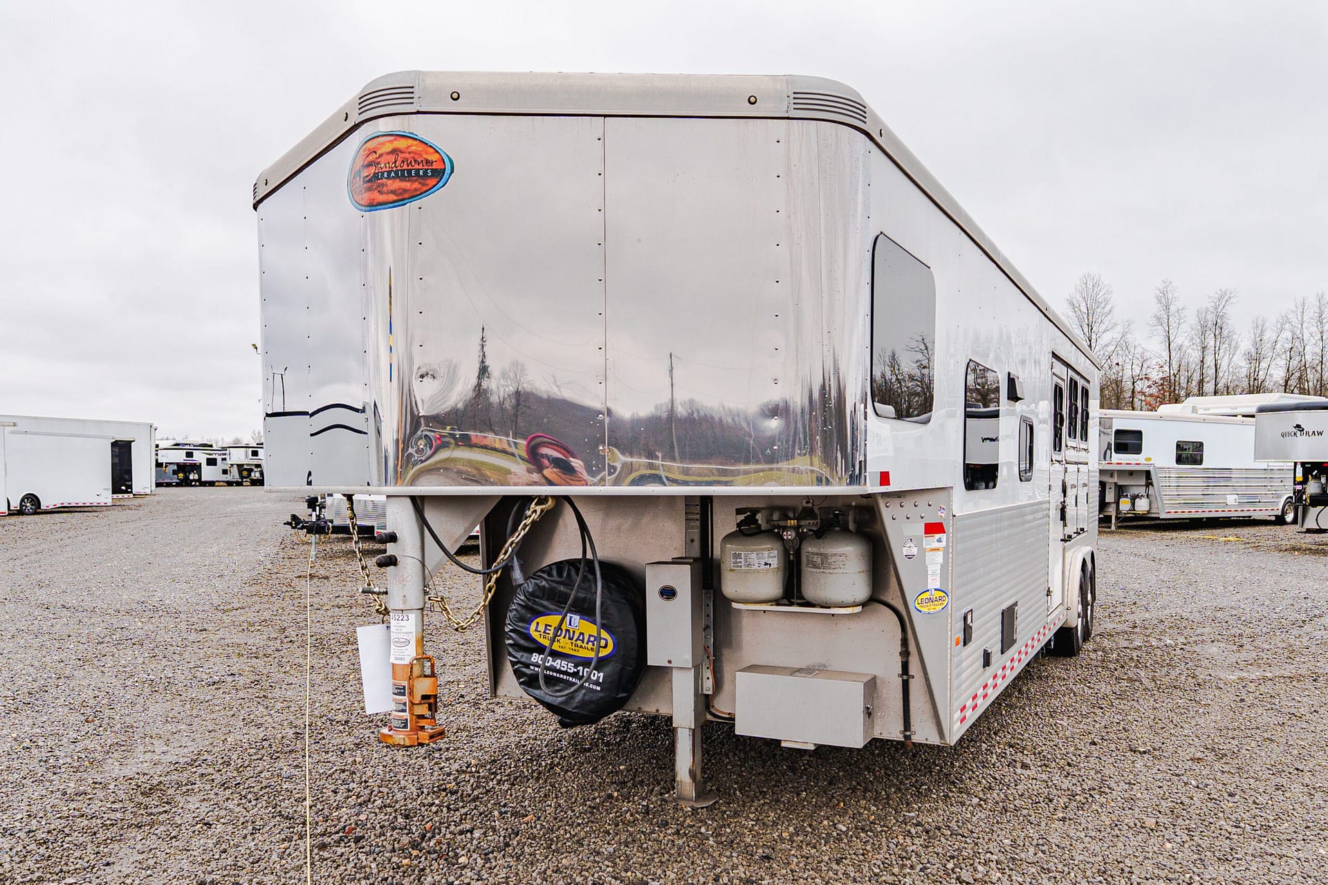 Shiny silver semi-trailer in a gravel lot, with a spare tire cover and propane tanks at the back, other trailers in the background.