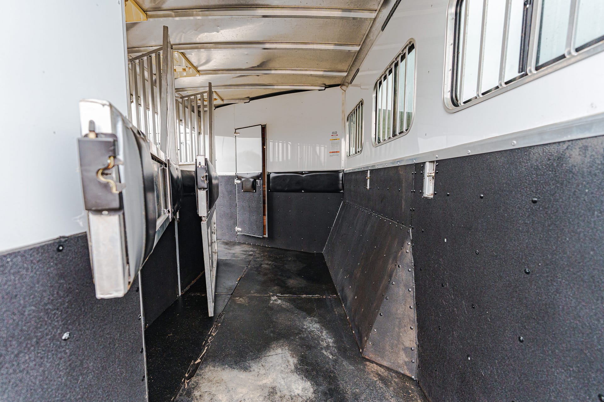 Interior of a horse trailer with metal divider bars along a narrow aisle and windows on the side walls, showing stall enclosures.