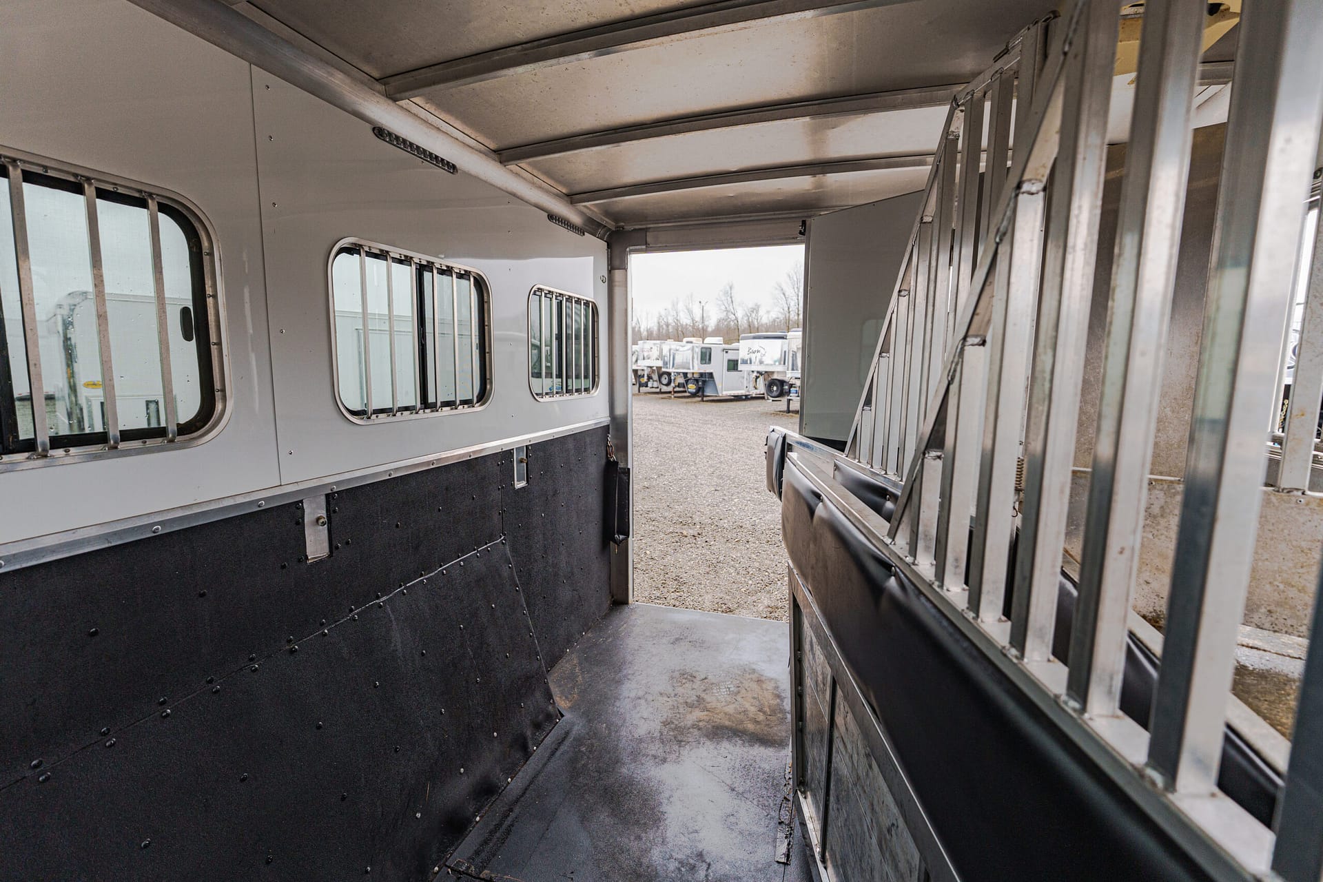 Interior of a metal livestock trailer with barred windows and a narrow walkway to an outdoor lot with other trailers visible outside.