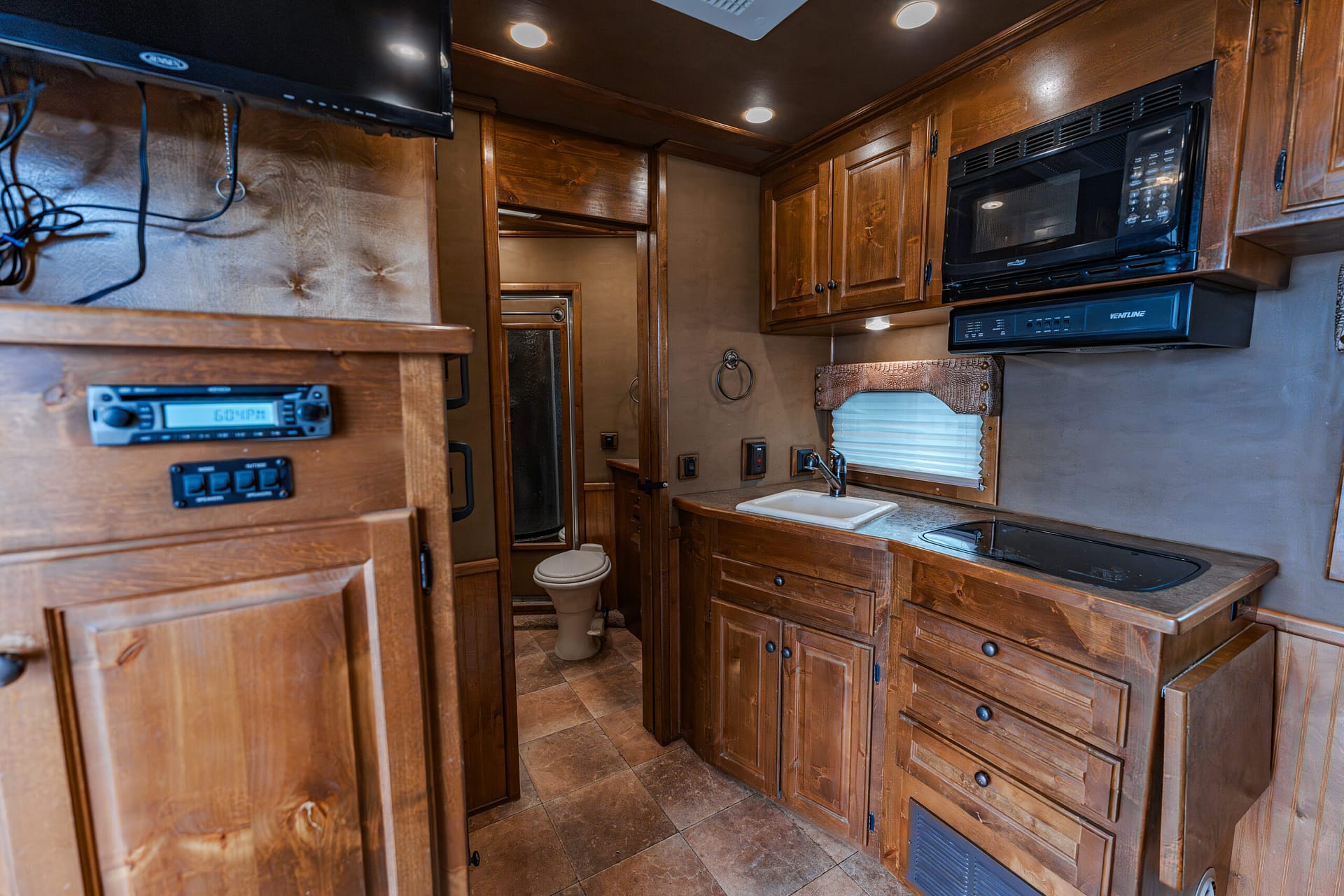 Wood-paneled RV kitchen with dark brown cabinets, a microwave and range hood above a counter, and a sink along a curved counter; a small hallway to a bathroom with a toilet is visible in the center. On the left, a mounted TV and control panel are seen; warm recessed ceiling lights illuminate the space.