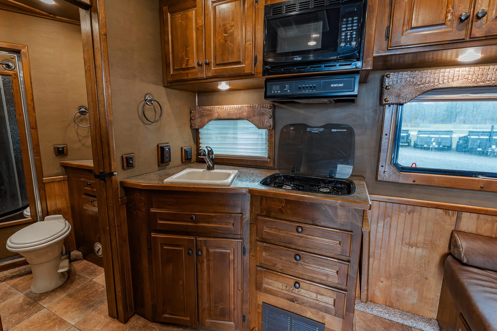 Wooden RV kitchen with microwave, stove, sink, and a window; built-in cabinets and drawers line the counter area.