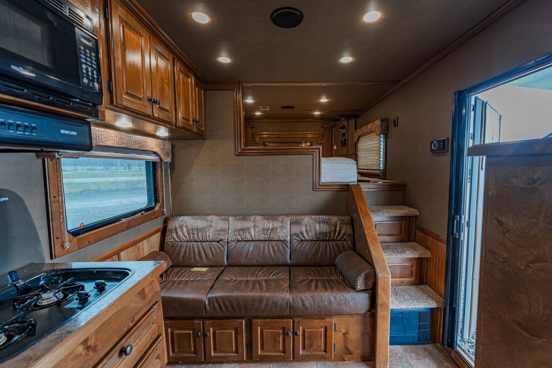 Interior of a wood-paneled RV living area featuring a brown leather sofa, upper cabinets, and a kitchenette along the left wall, with stairs and bedroom area toward the back.