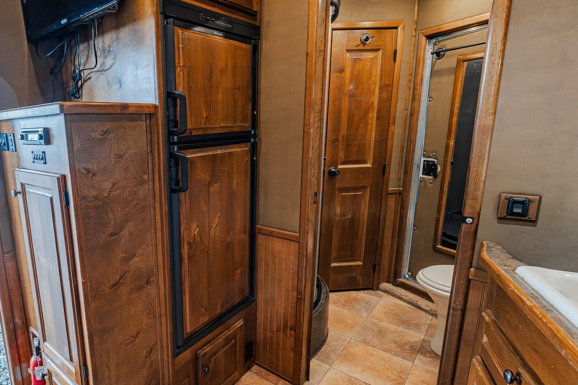 Rugged RV kitchen showing tall wooden cabinets, a black refrigerator, and a small door, with a toilet visible to the right side of the scene.