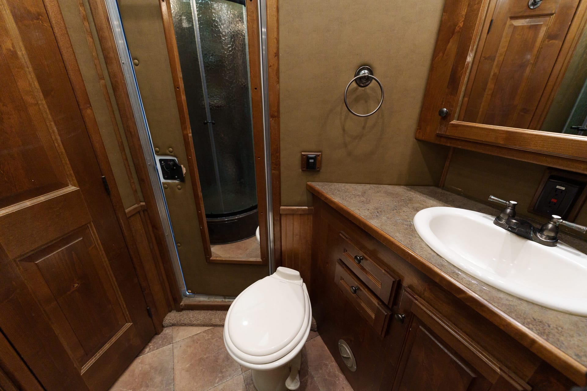 Wood-paneled bathroom with a white toilet in the foreground, adjacent sink and mirror cabinet along the right wall, and a glass shower stall on the left.