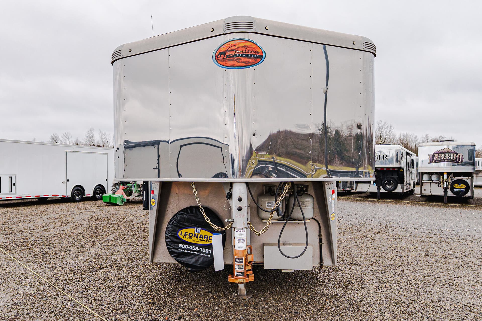 Front view of a large shiny aluminum trailer in a gravel lot, with a circular orange logo near the top and a Leonard Trailers spare-tire cover at the bottom left beneath the hitch.