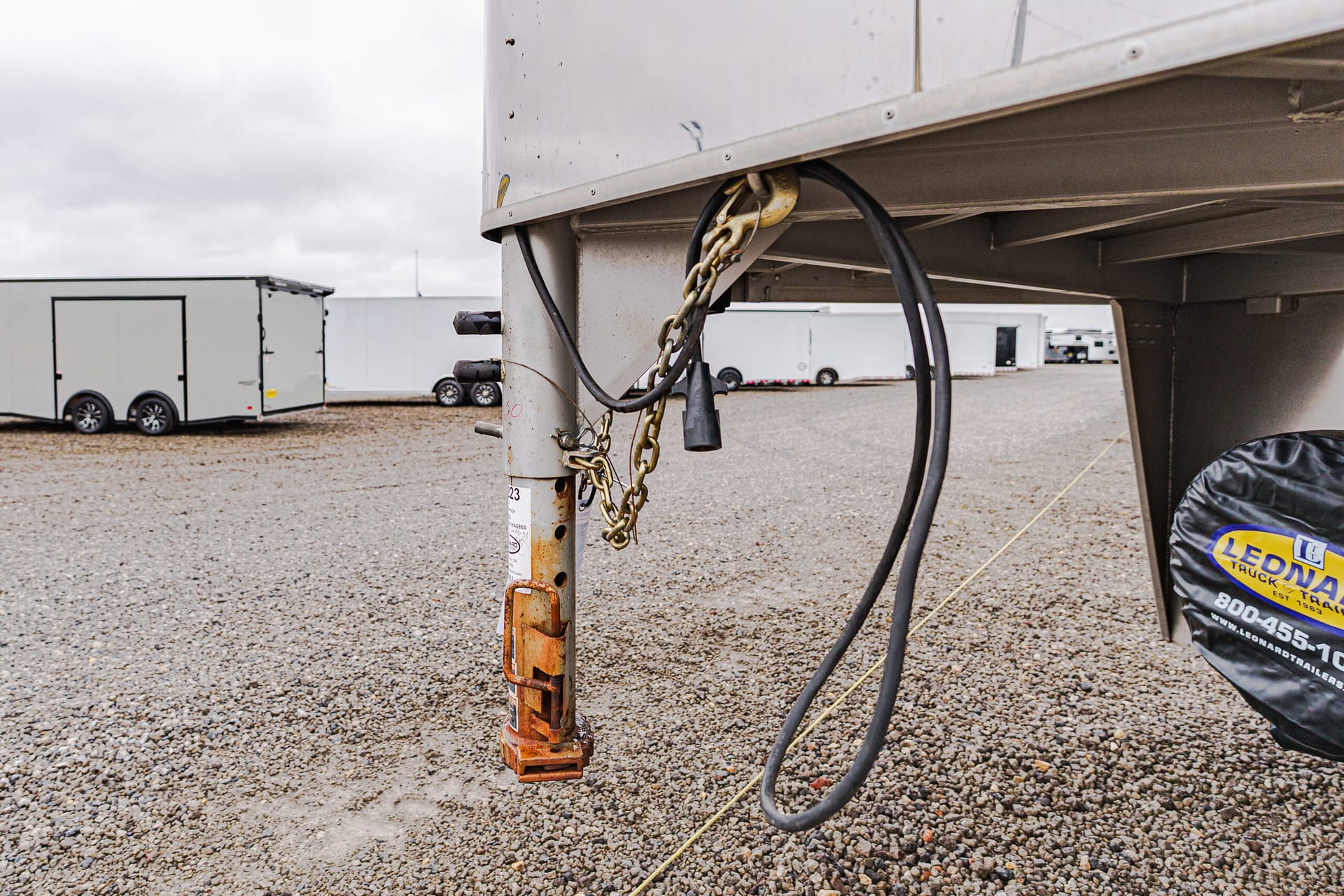 Close-up of a trailer hitch with rusted post, chains, and thick black cables in a gravel yard, with white trailers in the background.