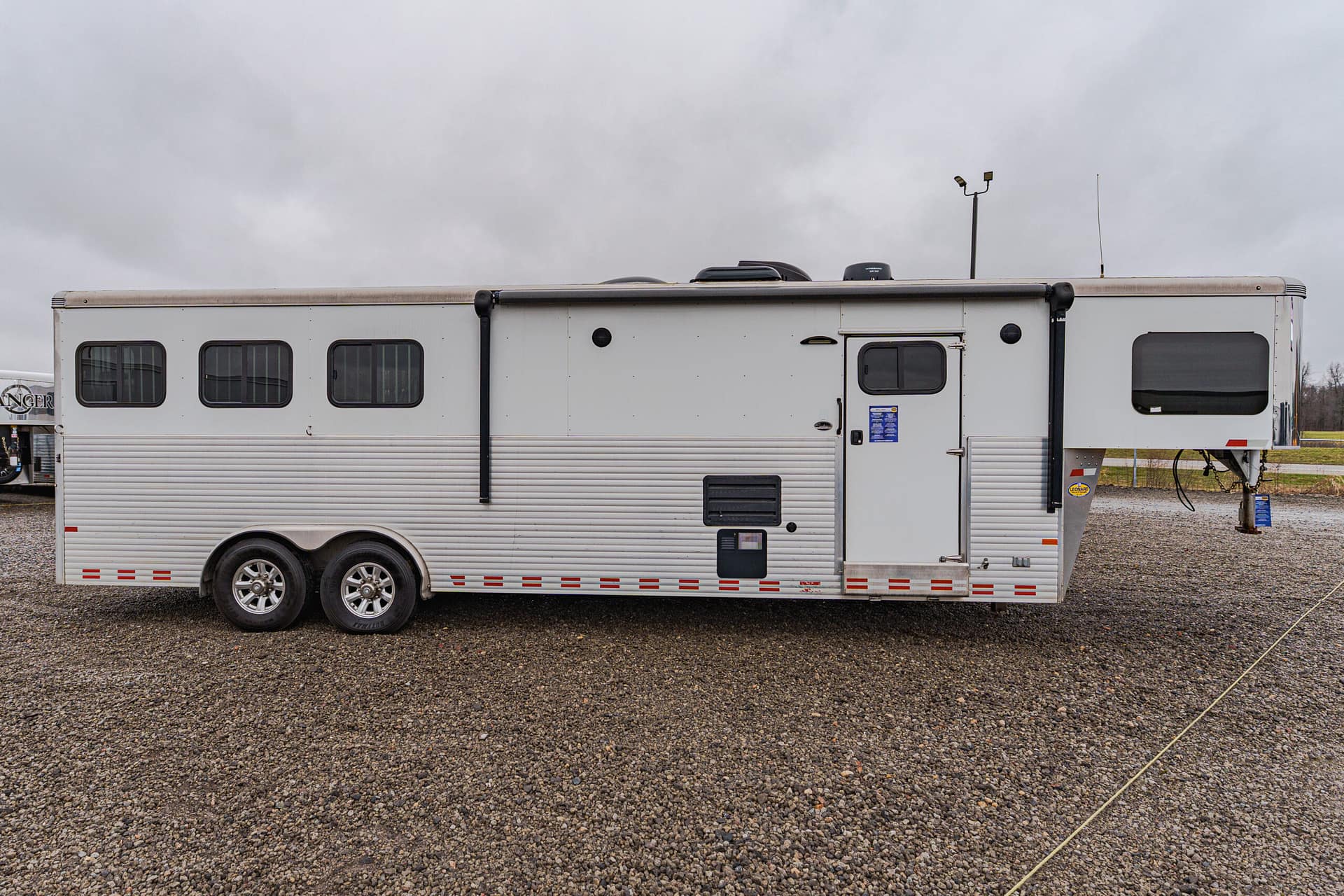 White travel trailer with multiple windows and a side door parked on a gravel lot under a cloudy sky.