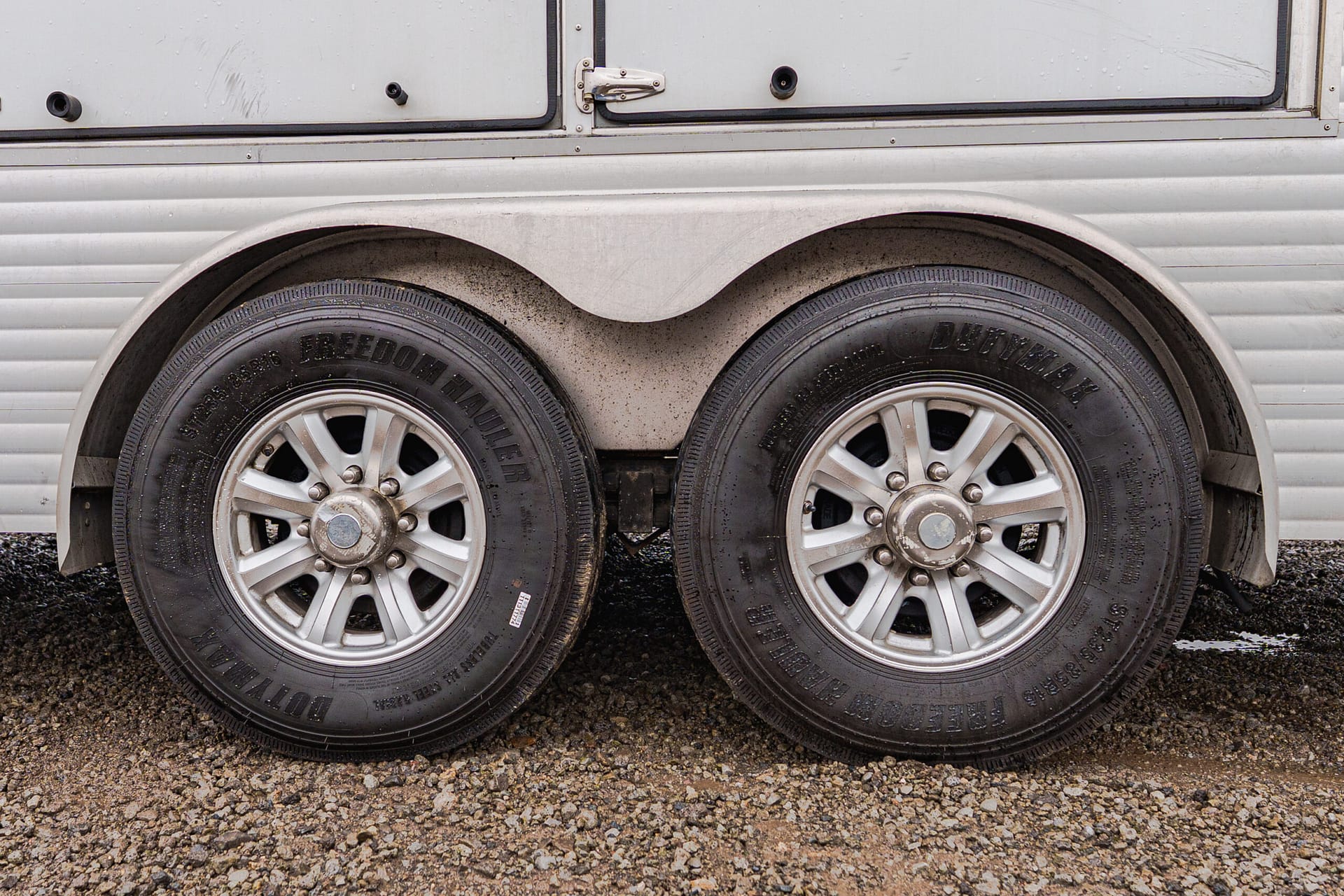 Close-up of a dual-axle trailer with two large tires and curved metal fender above on a gravel surface.
