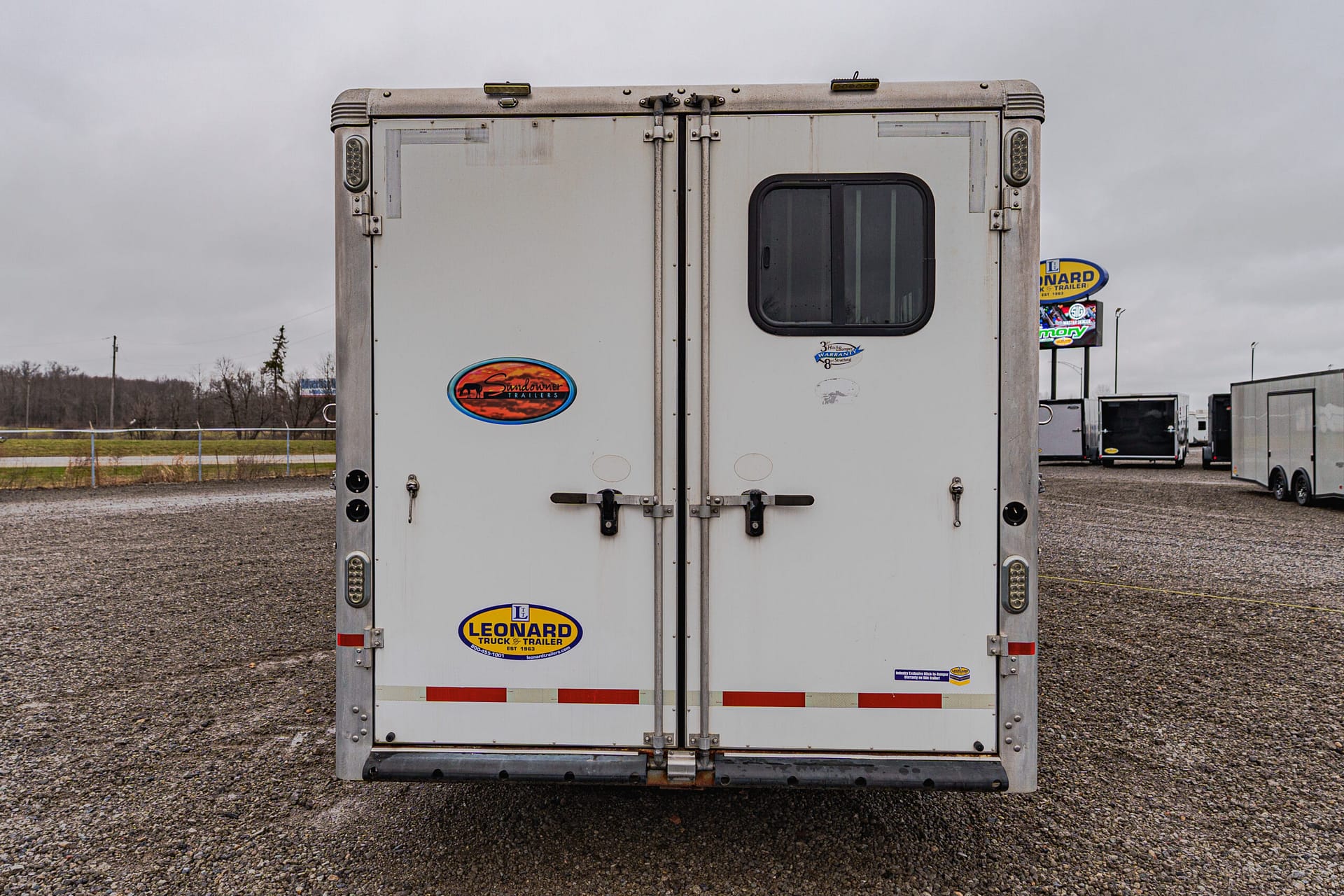 Back view of a large white trailer with double doors, a small window on the right door, and various brand stickers visible near the bottom in a gravel lot, with other trailers in the background.