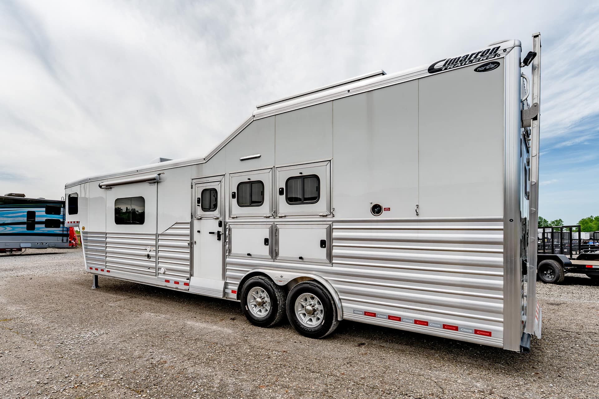 Silver livestock/utility trailer with multiple side doors and windows parked on a gravel lot under a cloudy sky