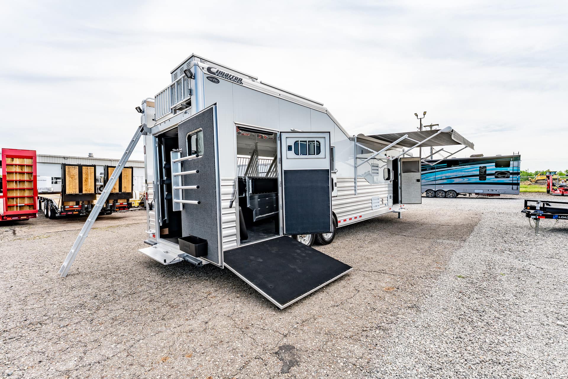 Silver utility trailer with open rear doors and extended ramp on a gravel lot, other trailers visible in the background.