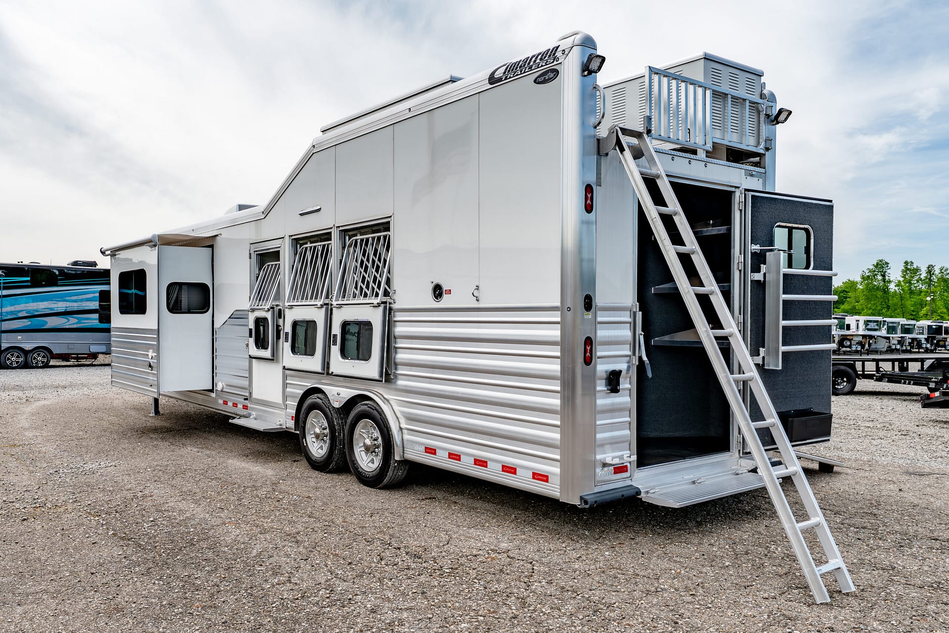 Silver aluminum trailer with multiple side windows and an exterior staircase at the rear, parked on a gravel lot with other trailers in the background.