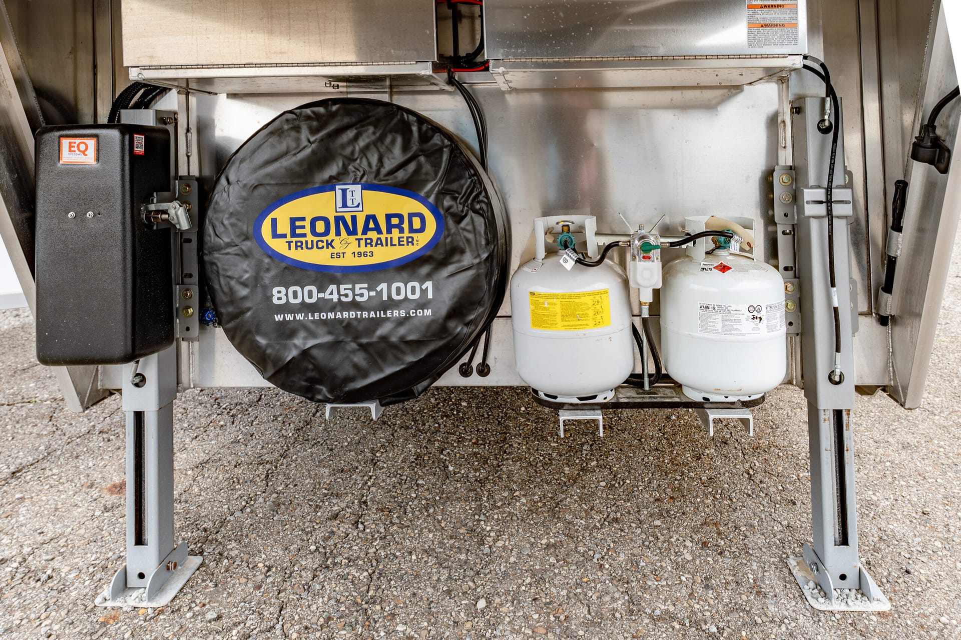 Underbelly of a trailer showing a spare tire with a Leonard Truck & Trailer cover and two propane tanks on a gravel surface