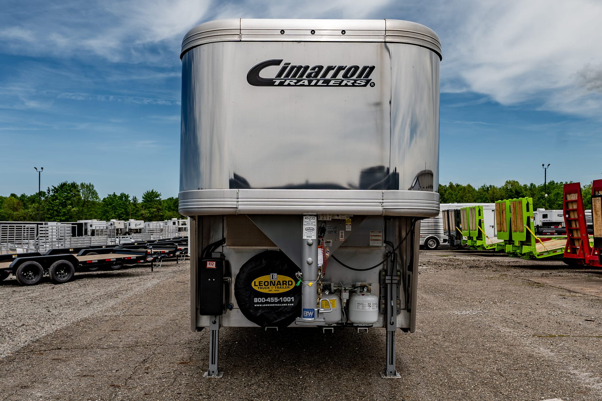 Rear view of a shiny Cimarron trailer in a lot with other trailers and equipment visible in the background, blue sky above.