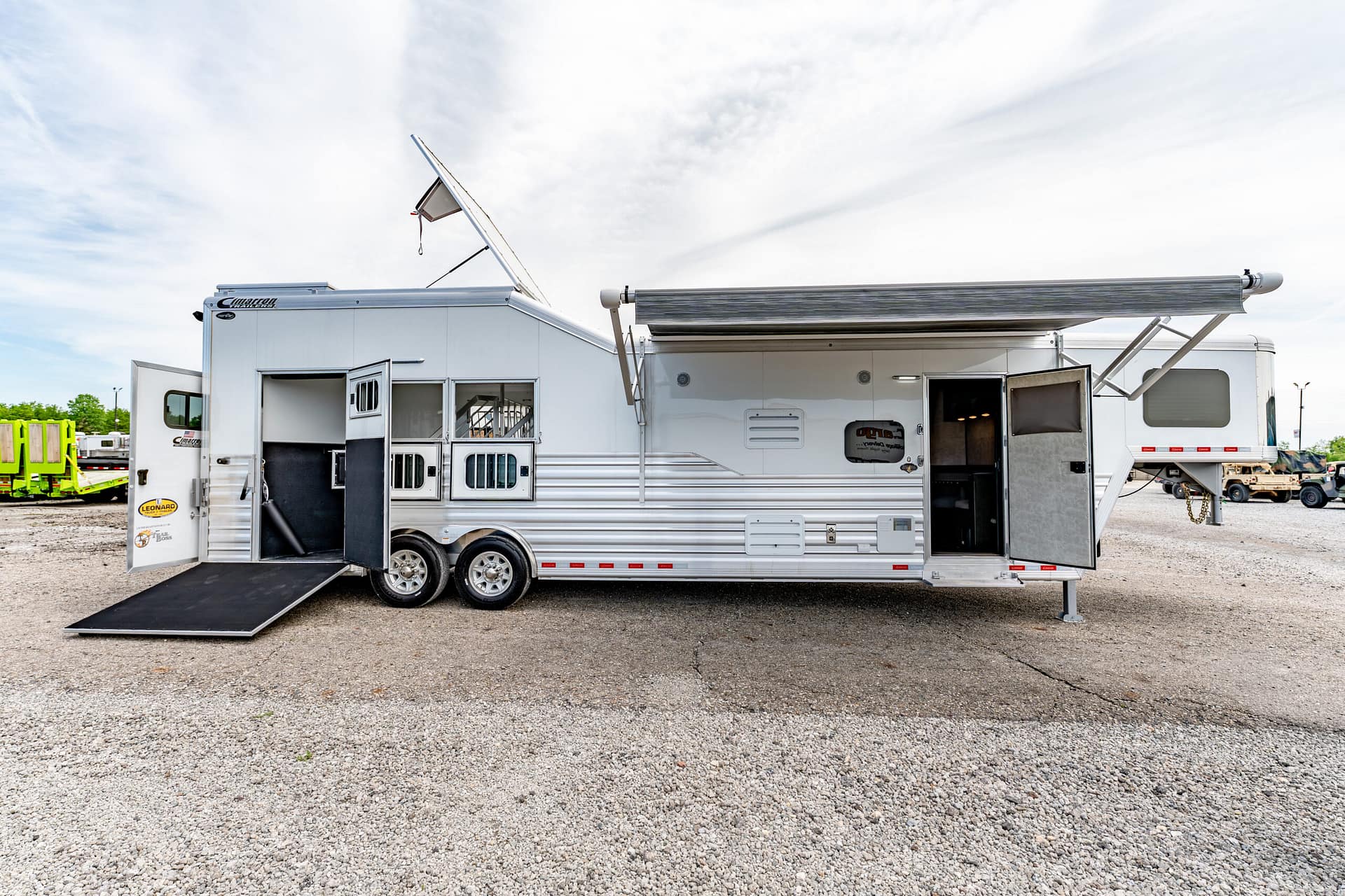 White horse trailer with a lowered ramp and open side door, parked on a gravel lot under a cloudy sky with an extended awning nearby.
