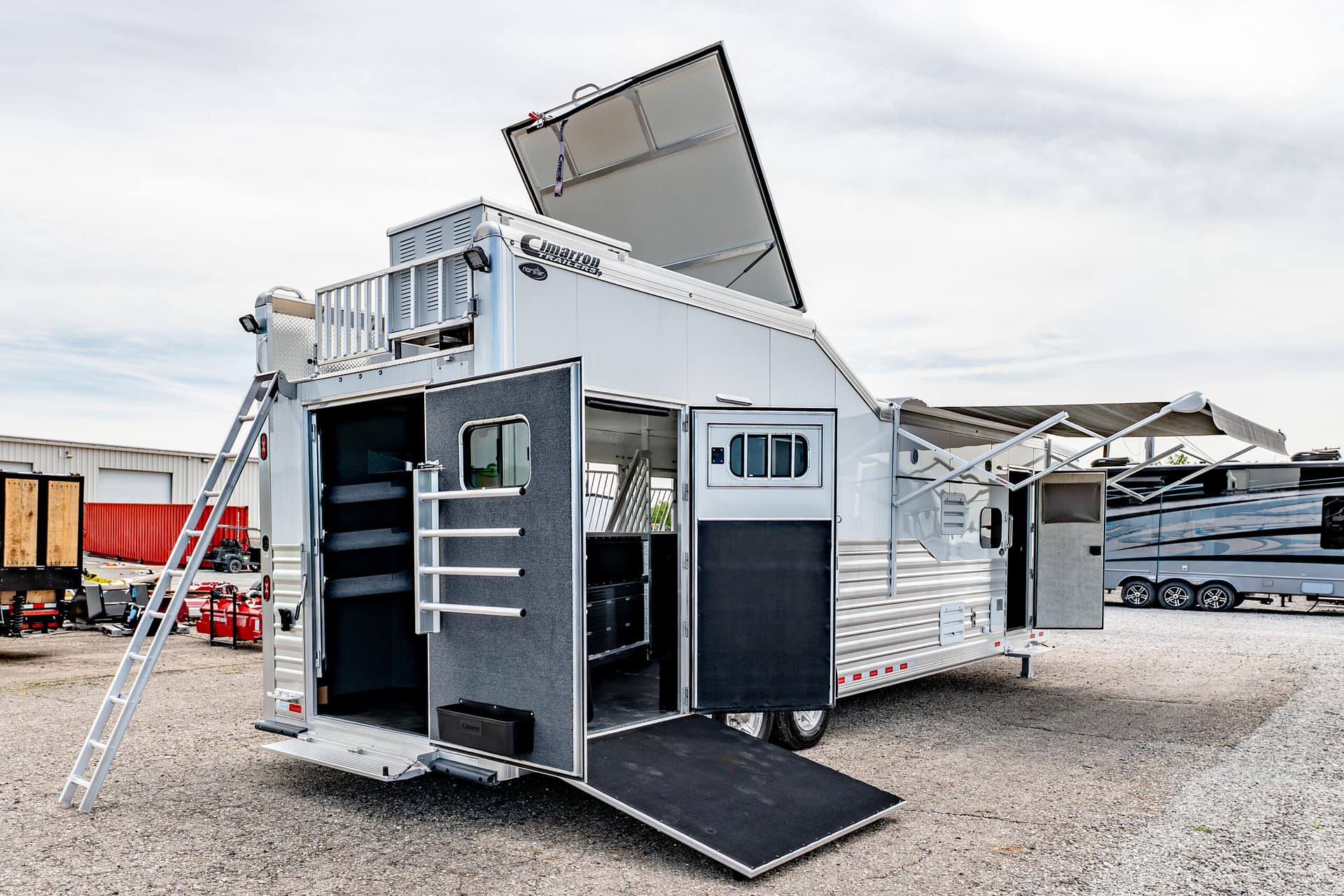Large silver aluminum trailer with its rear doors open and a ramp deployed on a gravel lot, outdoor stairs visible on the side