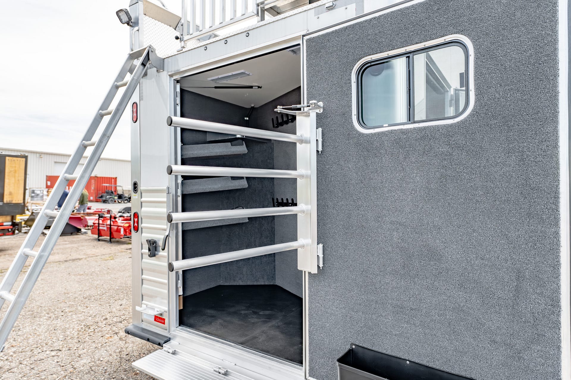 Rear of a gray textured trailer with its door open, showing a loading ramp and interior shelving in a gravel lot.