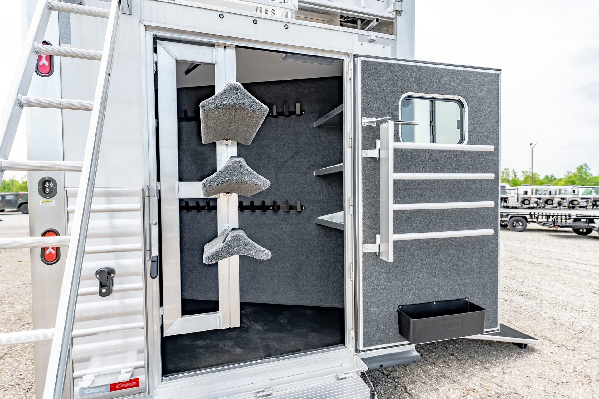 Open side entry of a silver aluminum trailer, showing a black padded interior with built-in racks and a ladder on the outside edge.