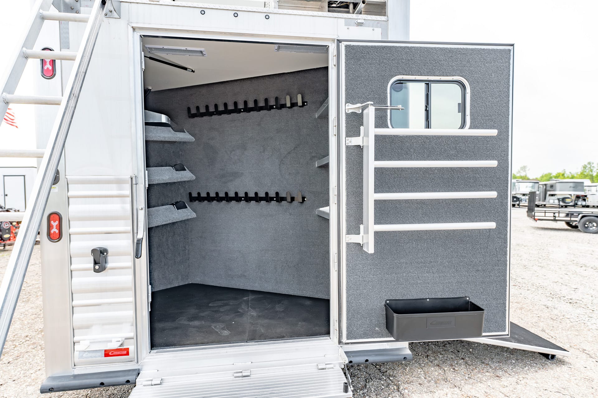 Open horse trailer rear with interior tack room and empty saddle racks along grey, carpeted walls plus exterior ladder visible on left side.
