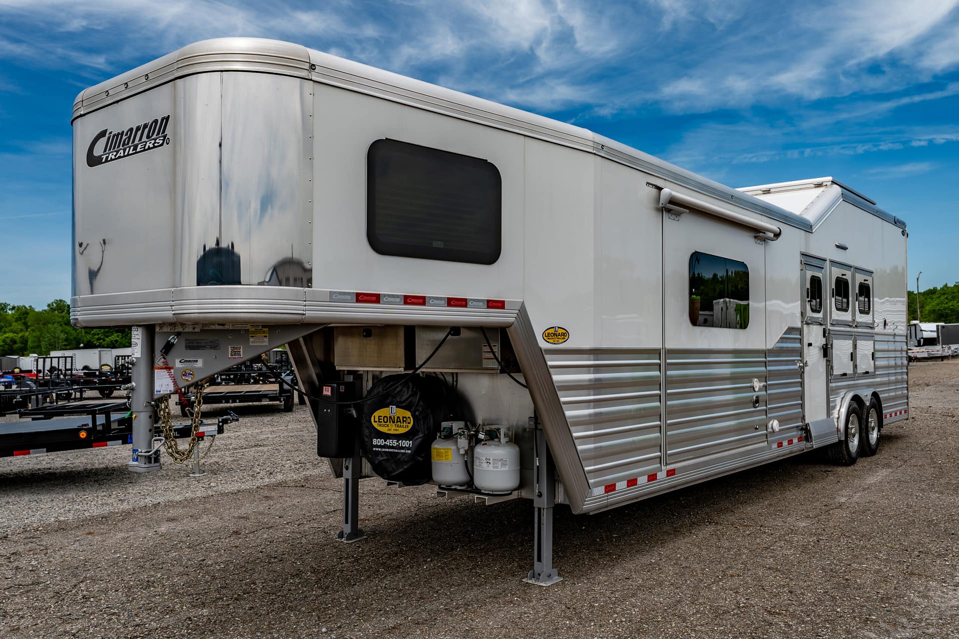 Silver travel trailer parked on gravel with a Cimarron Trailers logo and a blue sky backdrop, outdoors.