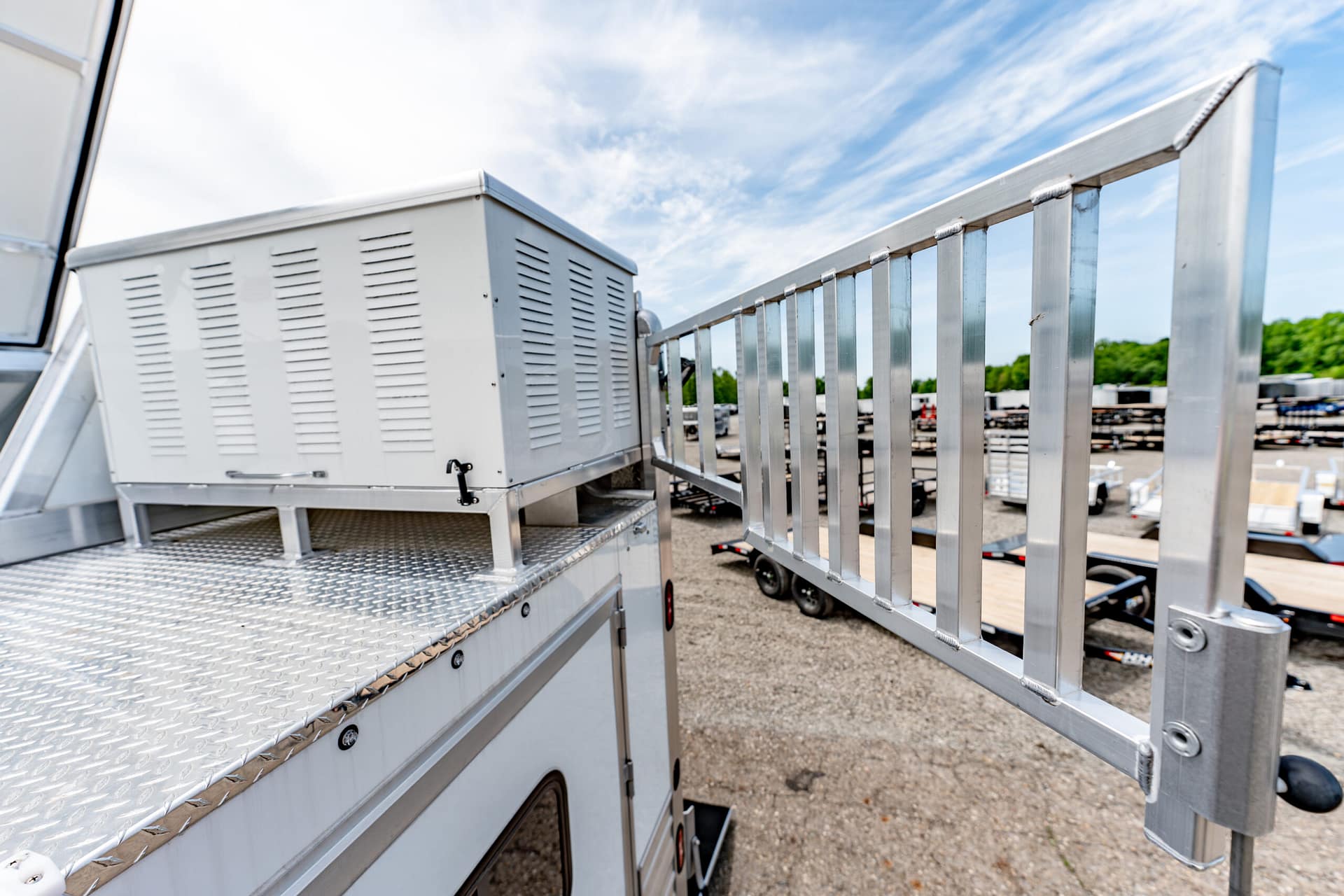 Aluminum utility trailer with a diamond-plate platform and vertical metal railing, parked in an outdoor yard under a blue sky.