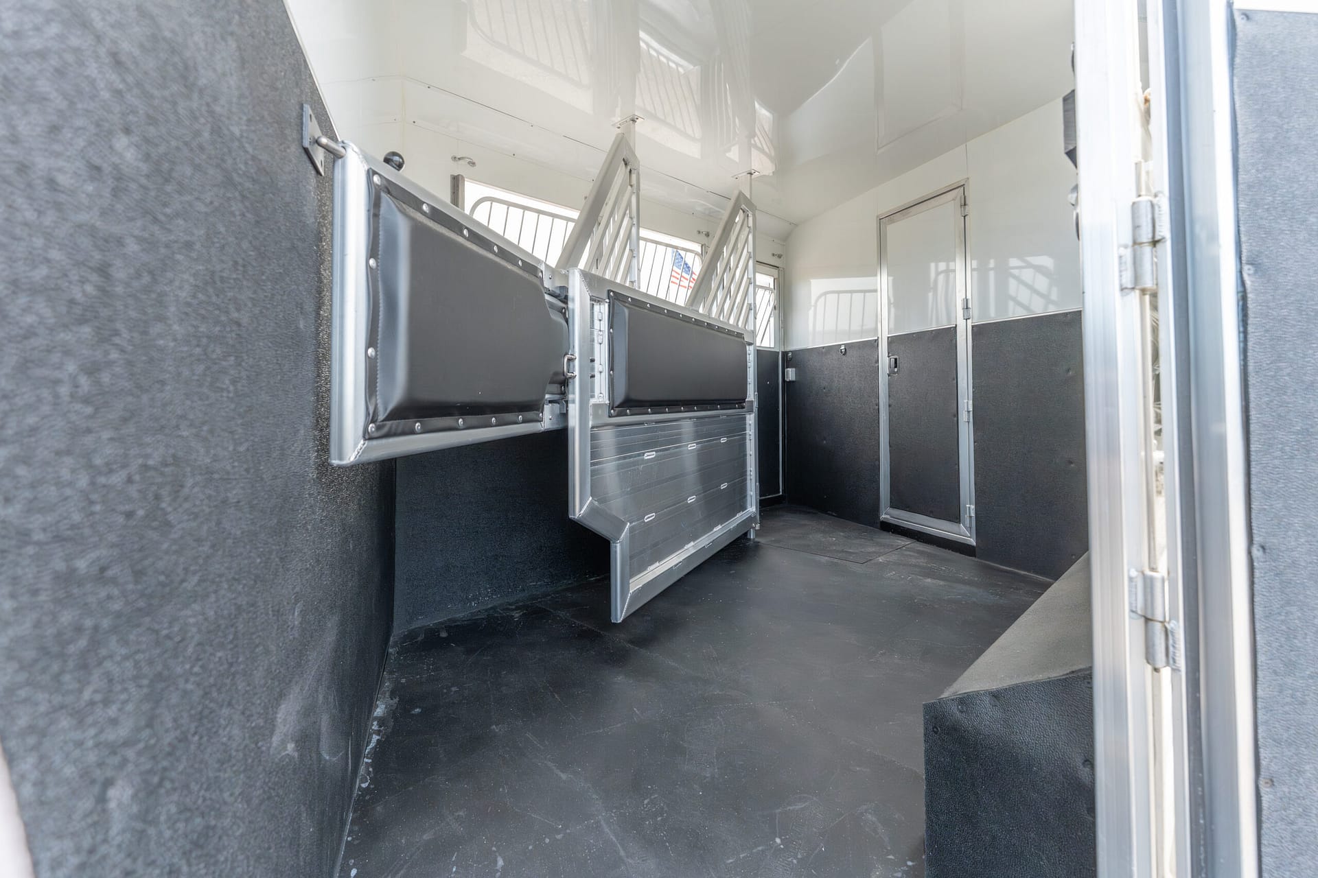 Interior of a horse trailer with metal dividing stalls and padded gray walls, empty and well-lit.