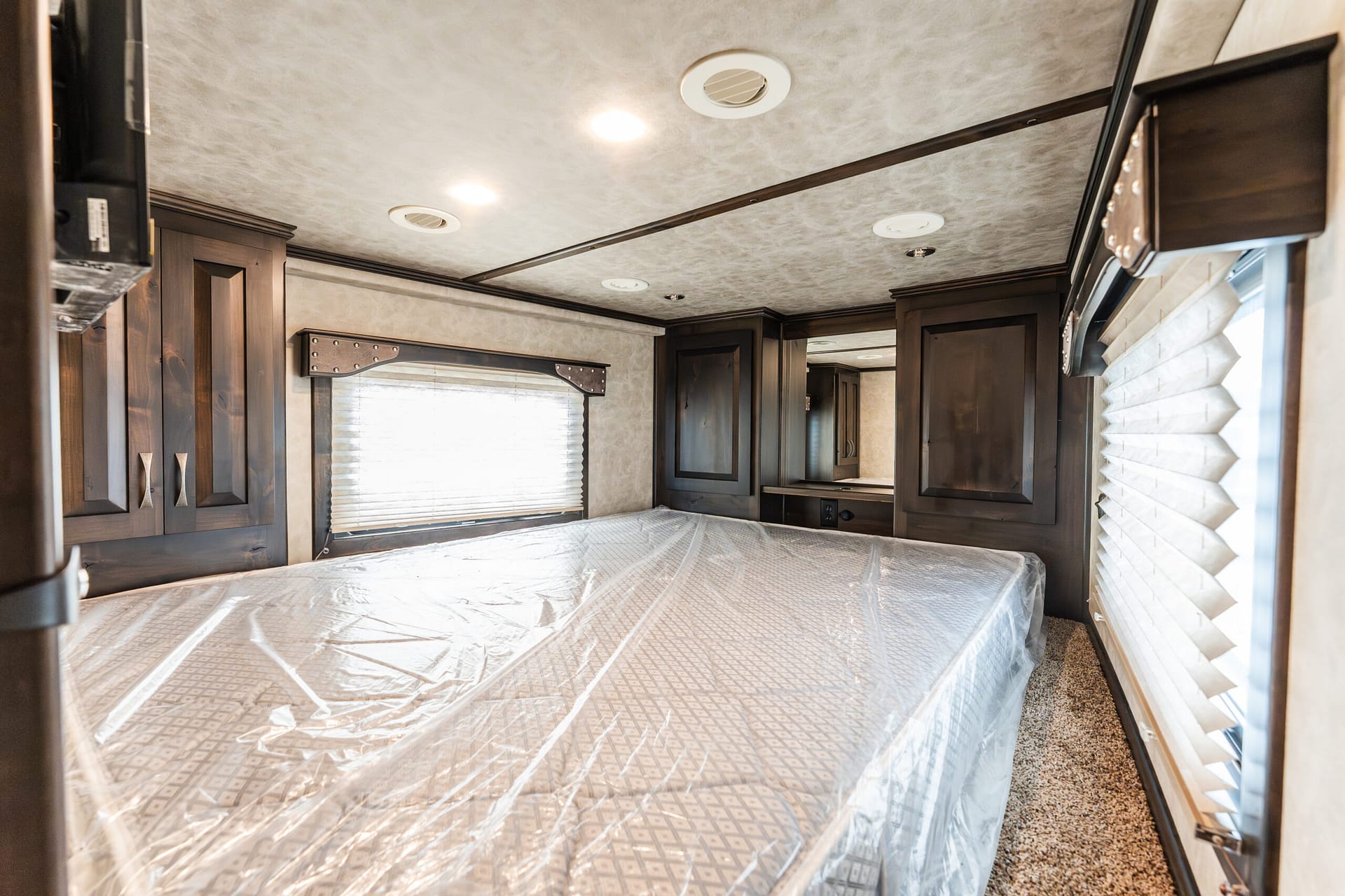 Wood-paneled RV bedroom with a large bed, dark cabinets, and built-in storage along the walls, window blinds on the left, and ceiling lights above.