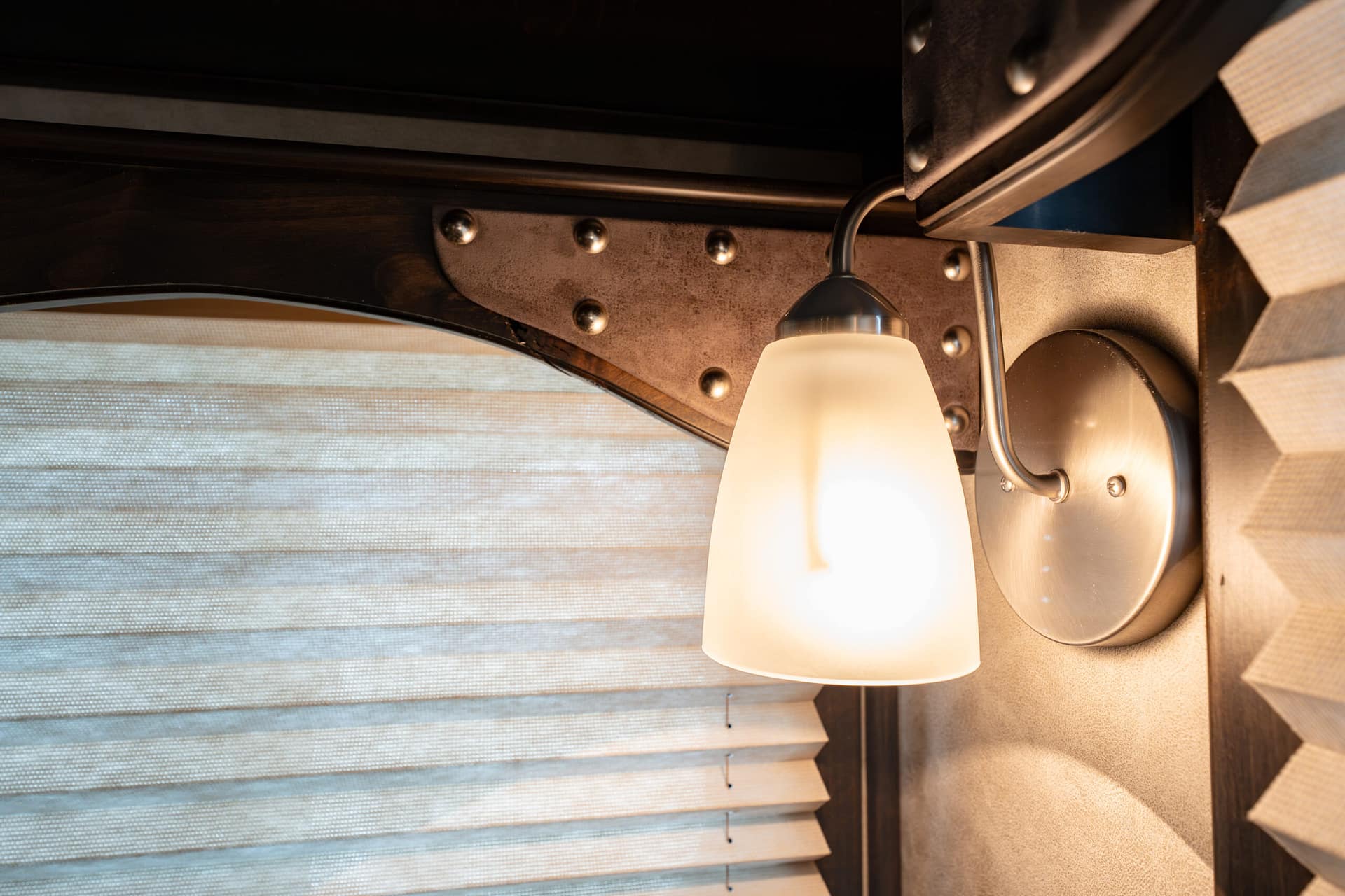 Close-up of a frosted glass lamp shade attached to a metal riveted plate beside horizontal window blinds.