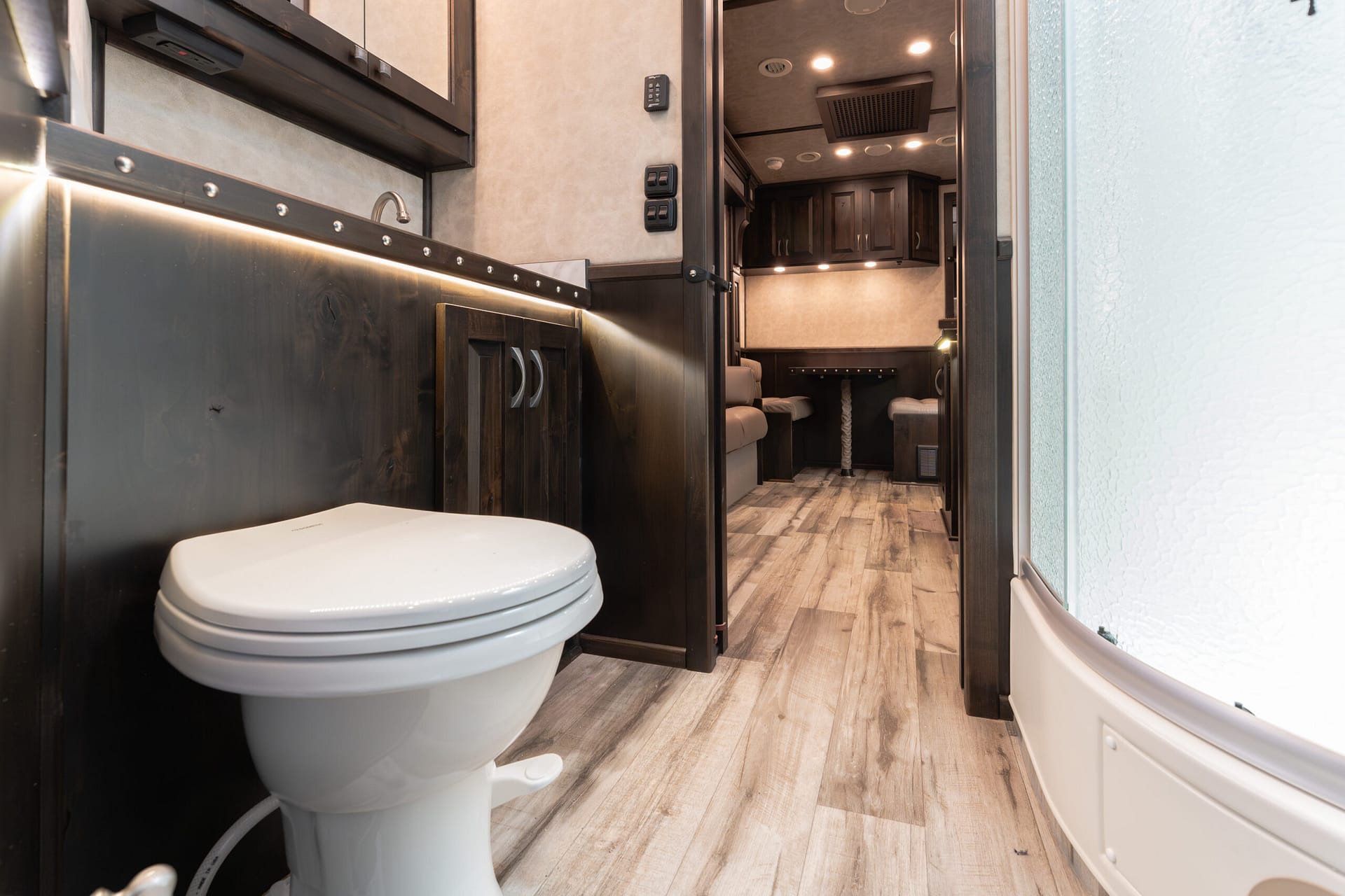 Interior shot of a compact RV bathroom: white toilet in the foreground, dark wood cabinets along the wall, and a doorway opening to a living area with seating and overhead lights.