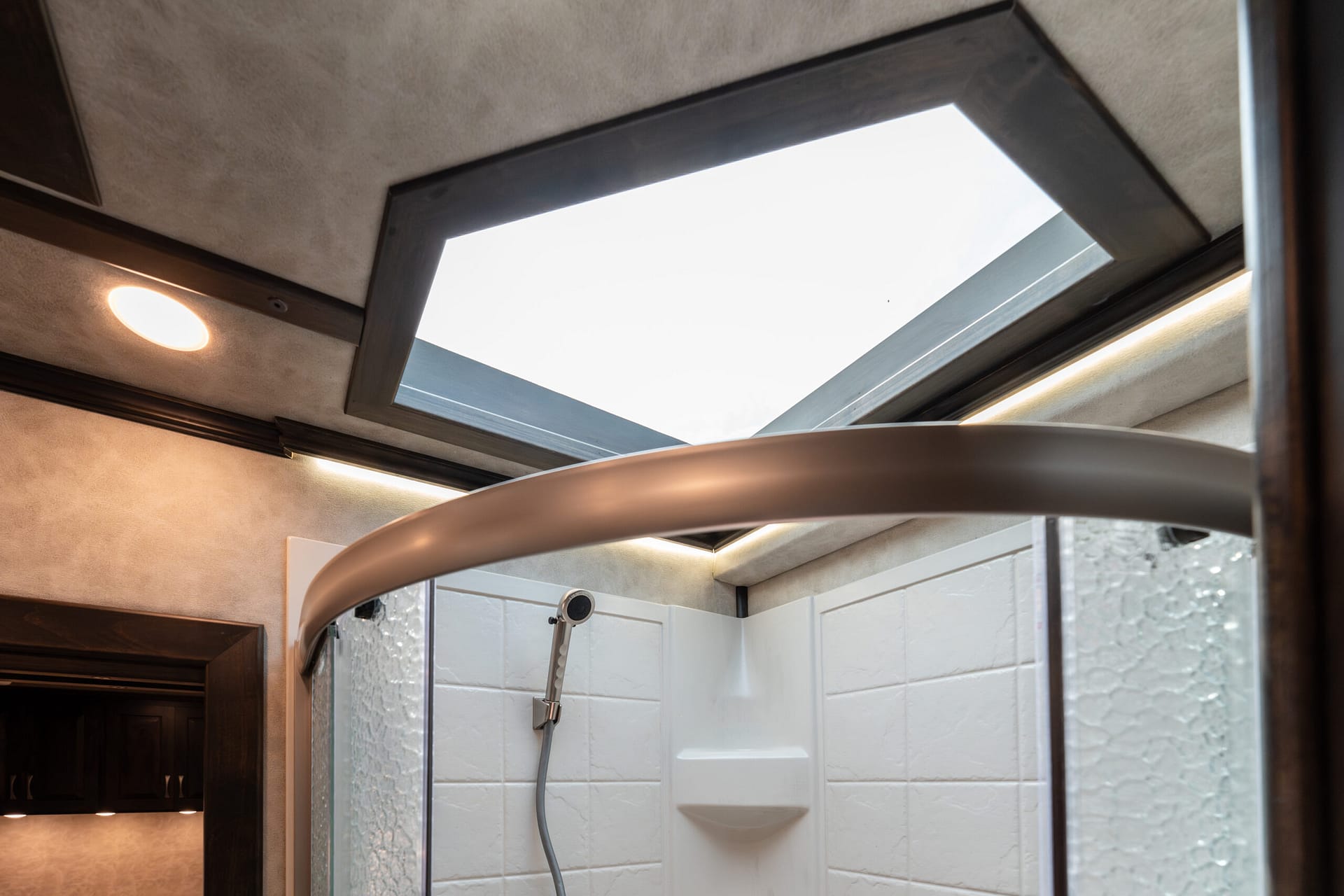 Shower stall with white tiled walls, glass doors, a curved metal bar, and a skylight bringing in daylight.