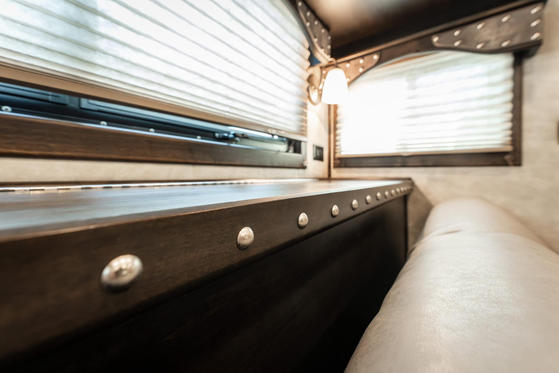 Interior of a mobile living space with dark wood cabinetry, riveted metal trim, and light beige seating beside striped blinds and warm lighting