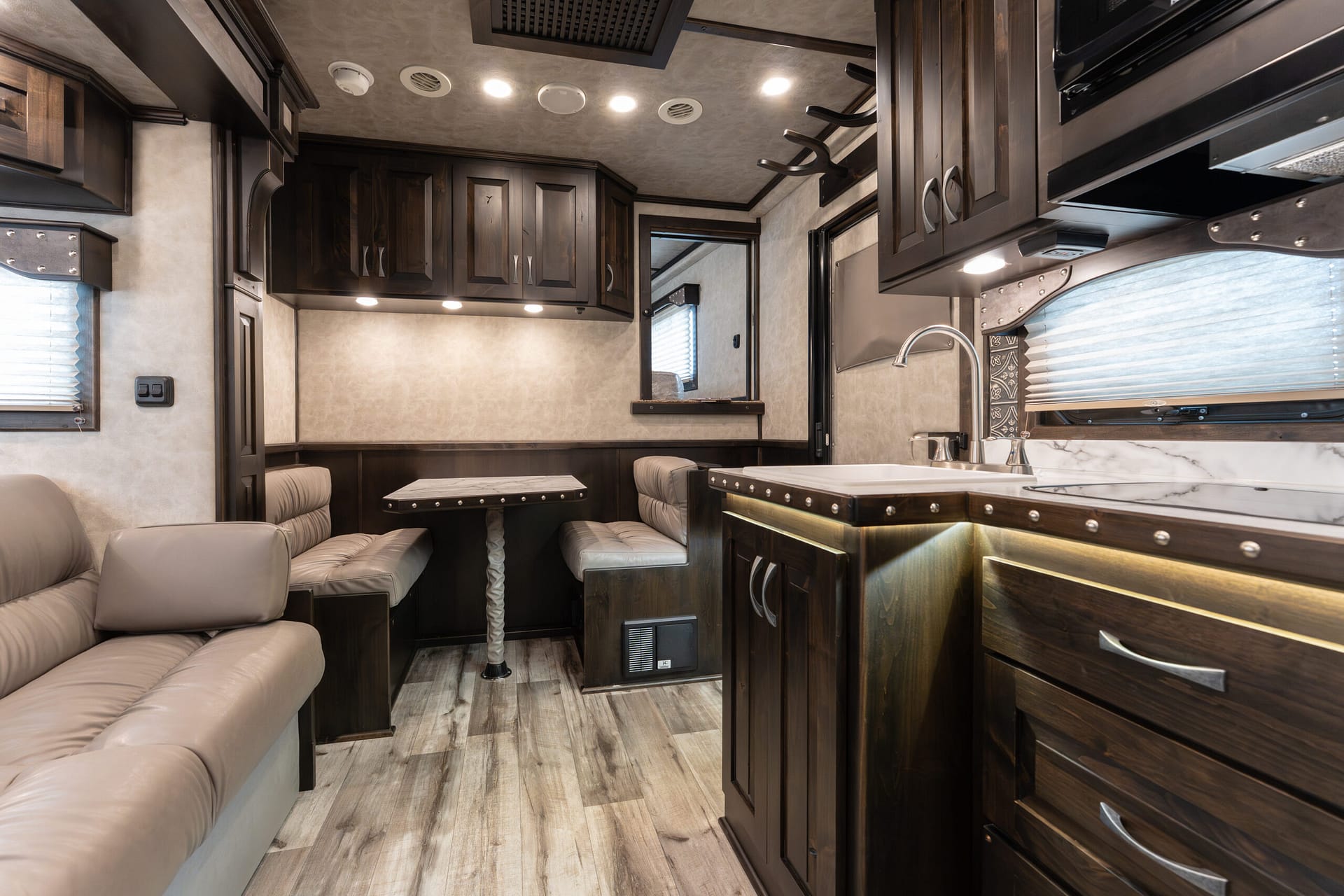 Interior of a luxury RV kitchen and living area with dark wood cabinets, beige leather seating, and a marble countertop.