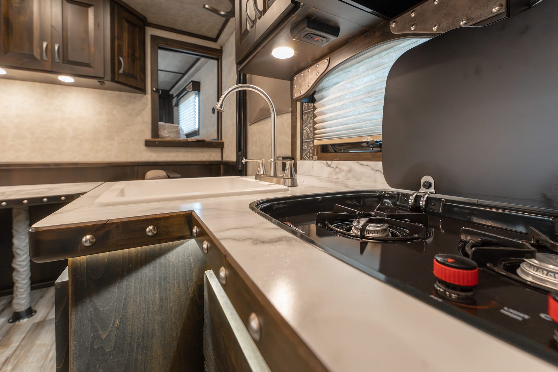 Interior of a compact RV kitchen with a stainless sink and gooseneck faucet, dark wood cabinets, and a propane stove with red control knob(s).