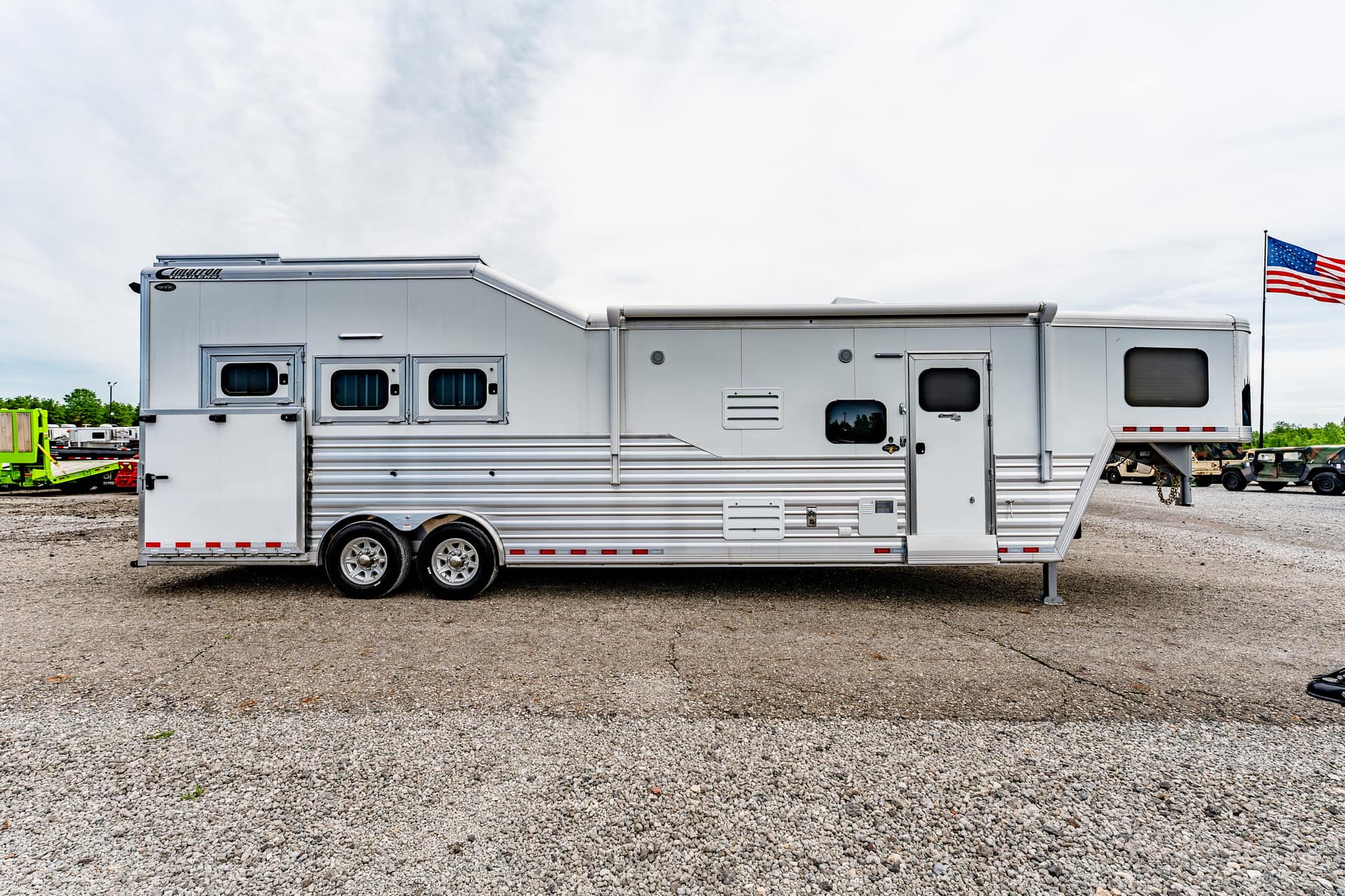 Long white travel trailer on a gravel lot with an American flag fluttering in the distance.