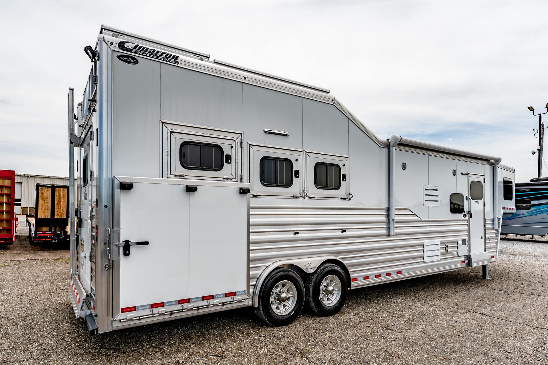 Long silver horse trailer with two side windows and chrome trim, parked on a gravel lot.