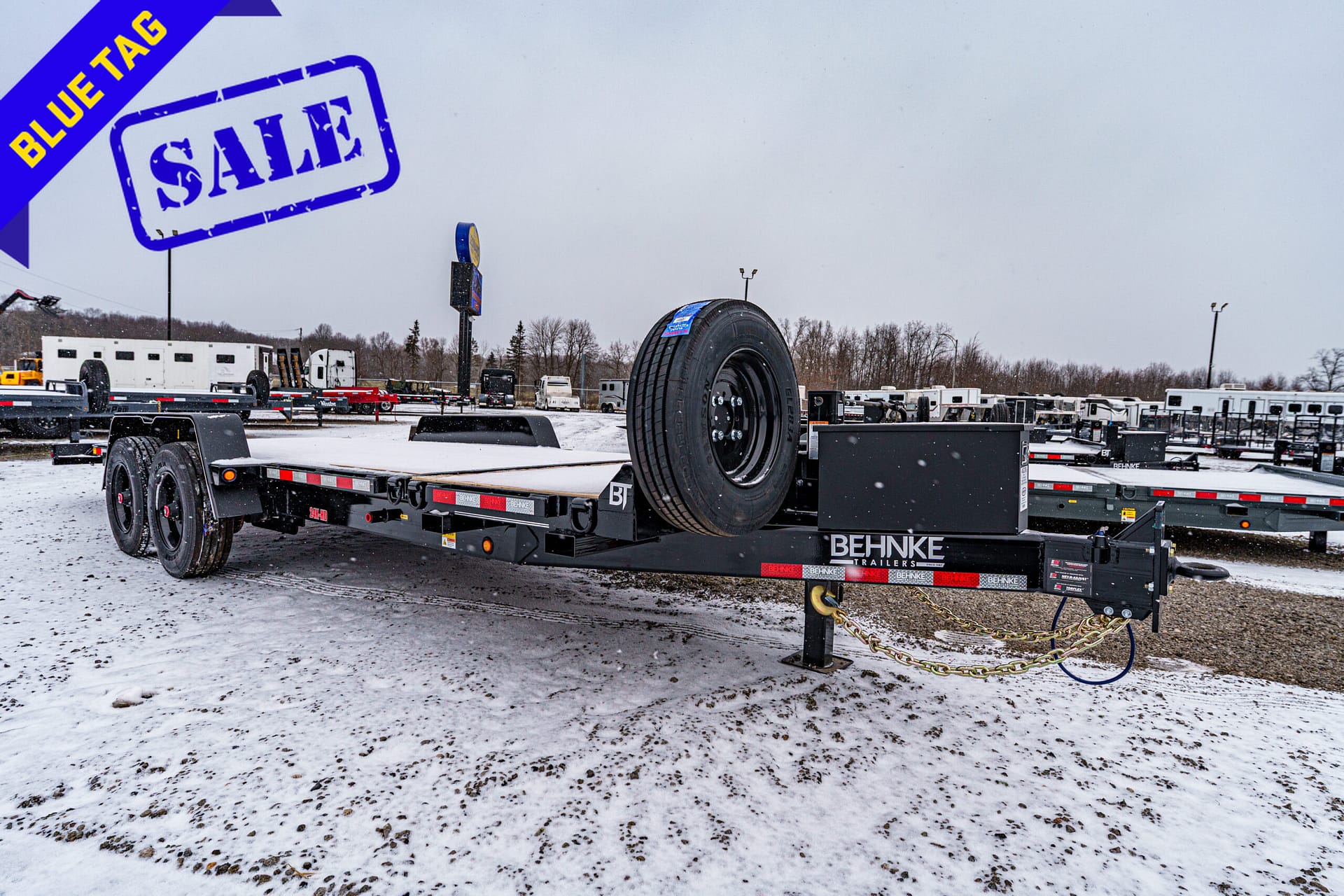 Blue sale banner overlays a snowy trailer lot, with a flatbed trailer carrying a spare tire in the foreground and rows of trailers in the background.