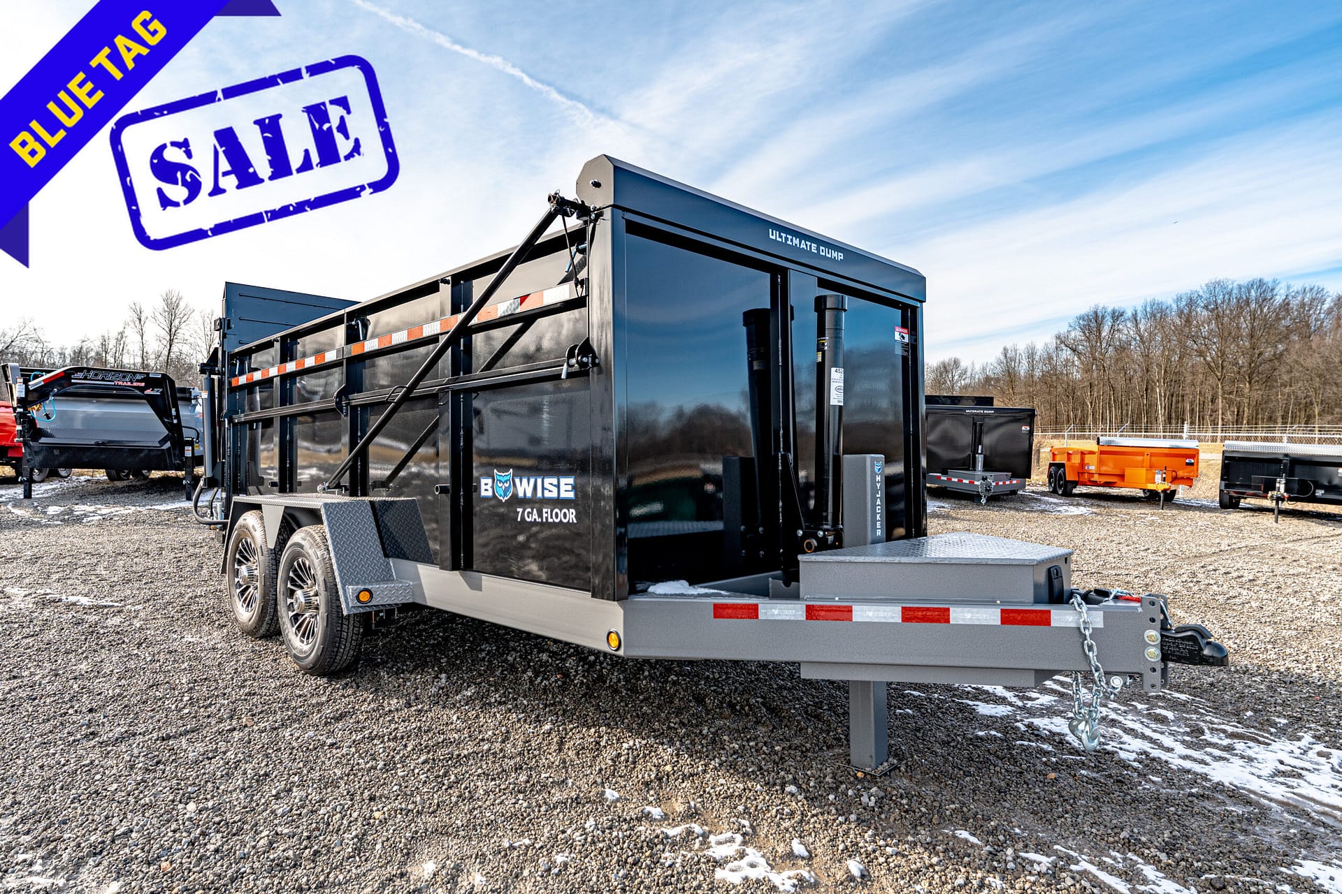 Black Ultimate Dump trailer on a gravel lot, with a blue 'Blue Tag Sale' banner in the top left corner and light snow patches on the ground.