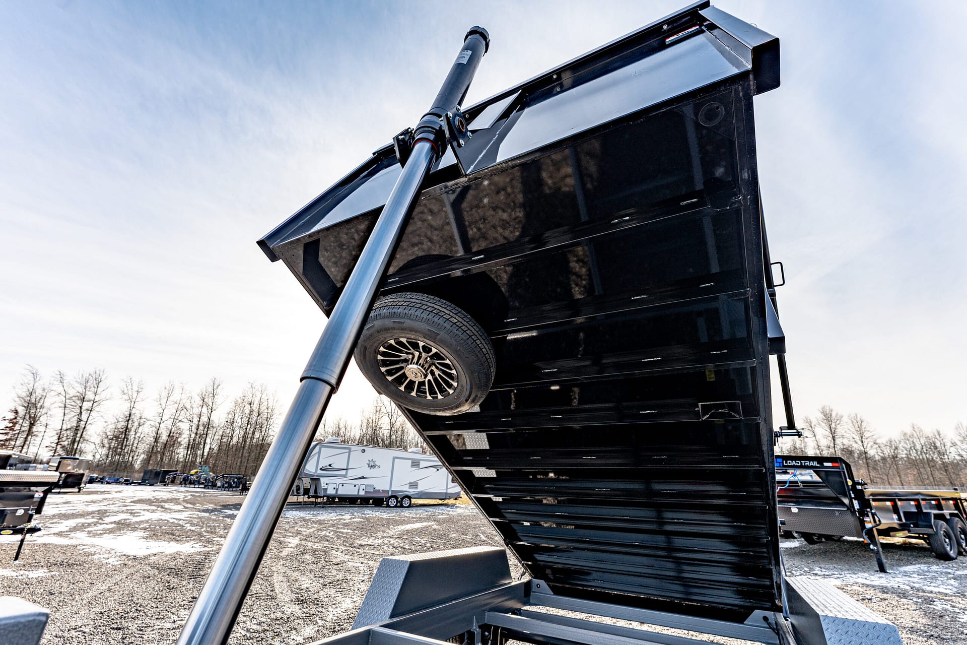 Black dump trailer tilted upward by a hydraulic lift in a dealership lot, with other trailers visible in the background as a blue sky overhead.