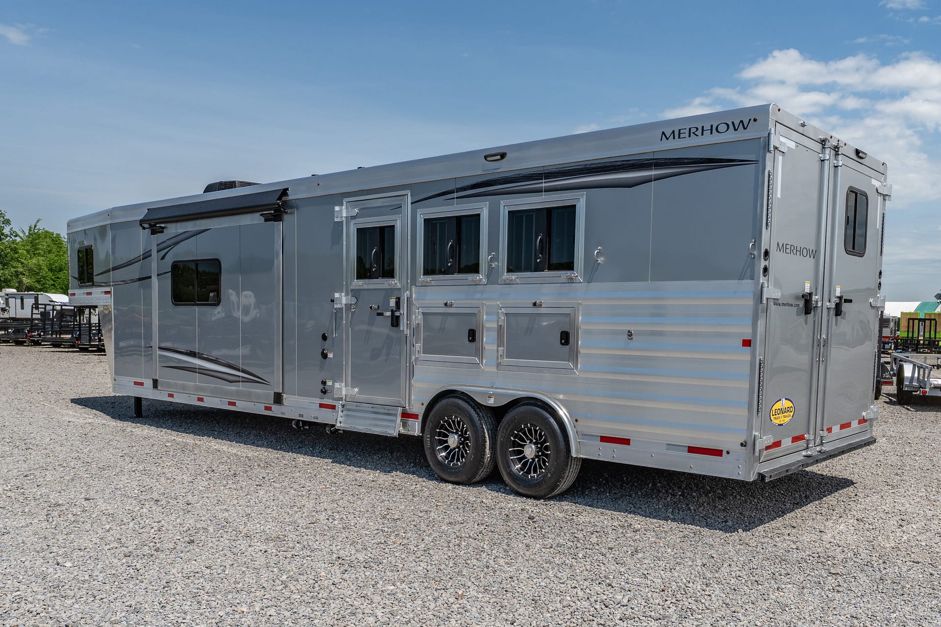 Silver Merhow horse trailer with multiple side windows and tandem wheels parked on a gravel lot under a blue sky
