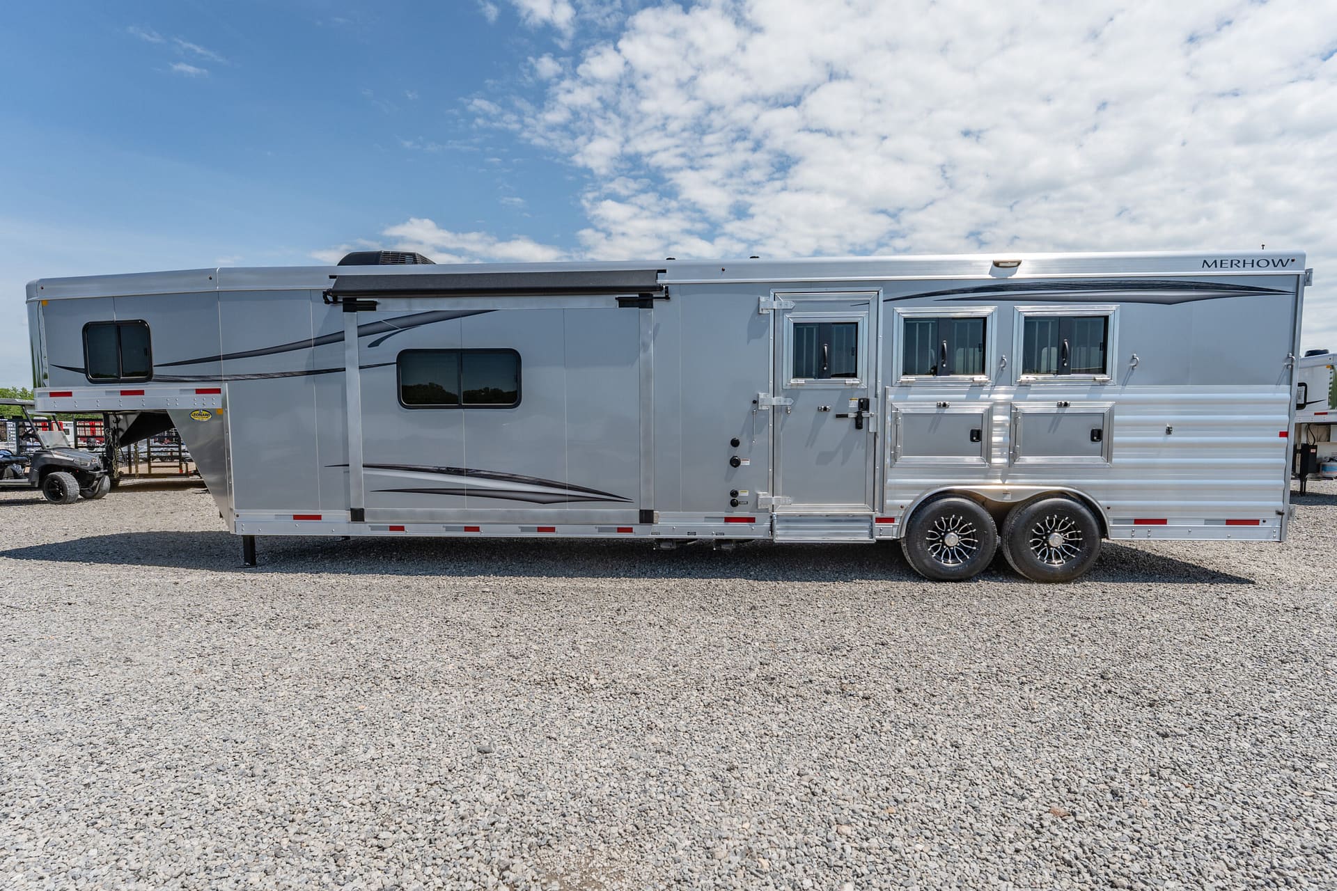 Silver Merhow horse trailer seen from the side, with multiple windows and storage compartments on a gravel lot under a blue sky.