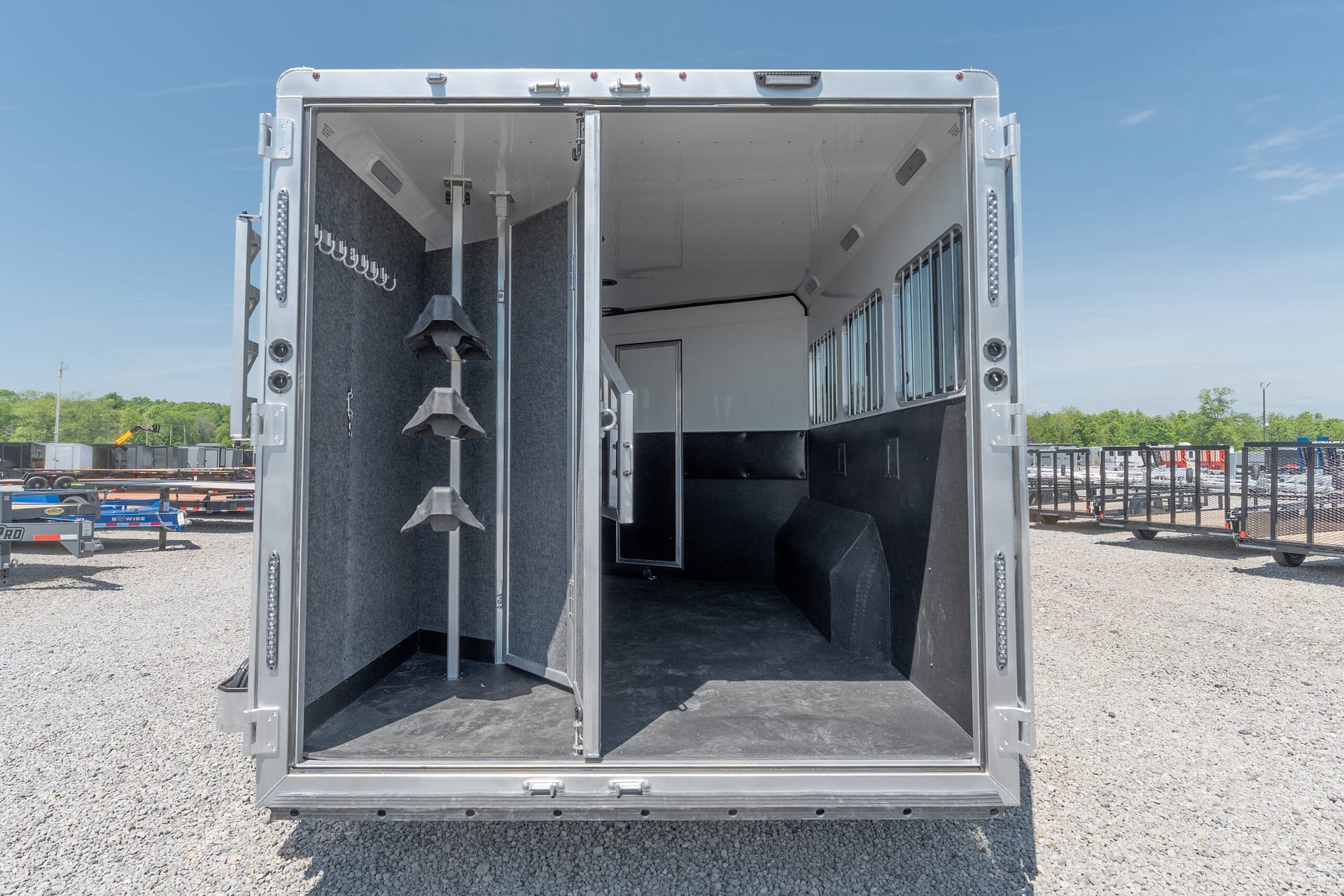 Open horse trailer showing tack hooks and partitioned stalls inside, parked on a gravel lot with other trailers in the background.