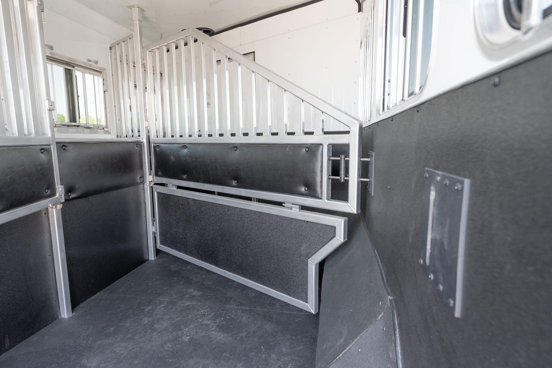 Interior of a metal horse trailer with dividing rails and padded lower walls, viewed from the aisle edge.