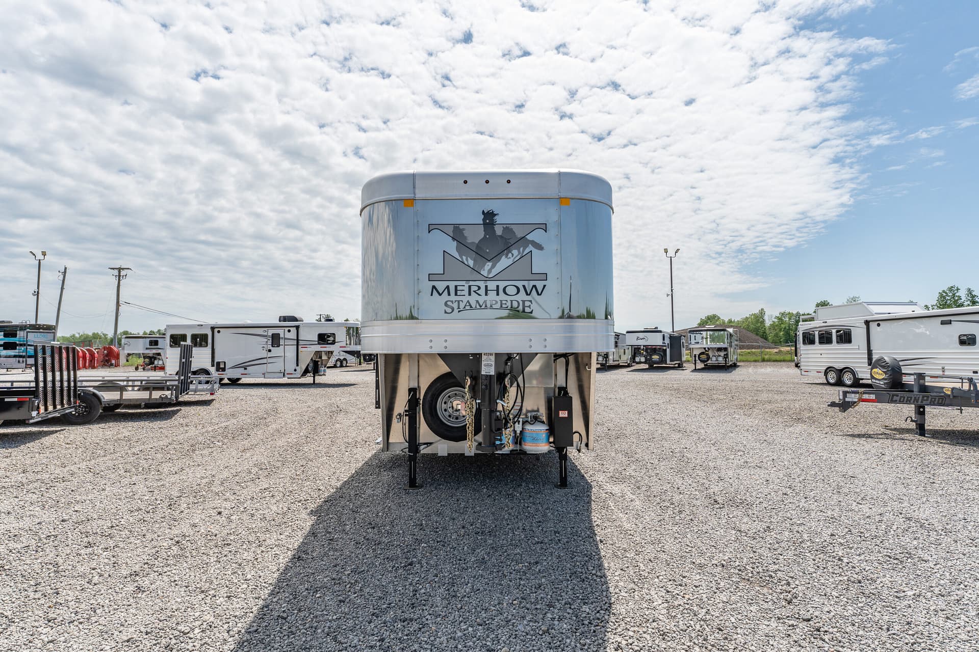 Front view of a large, shiny Merhow Stampede horse trailer parked in a gravel lot with other trailers and a cloudy blue sky behind it.