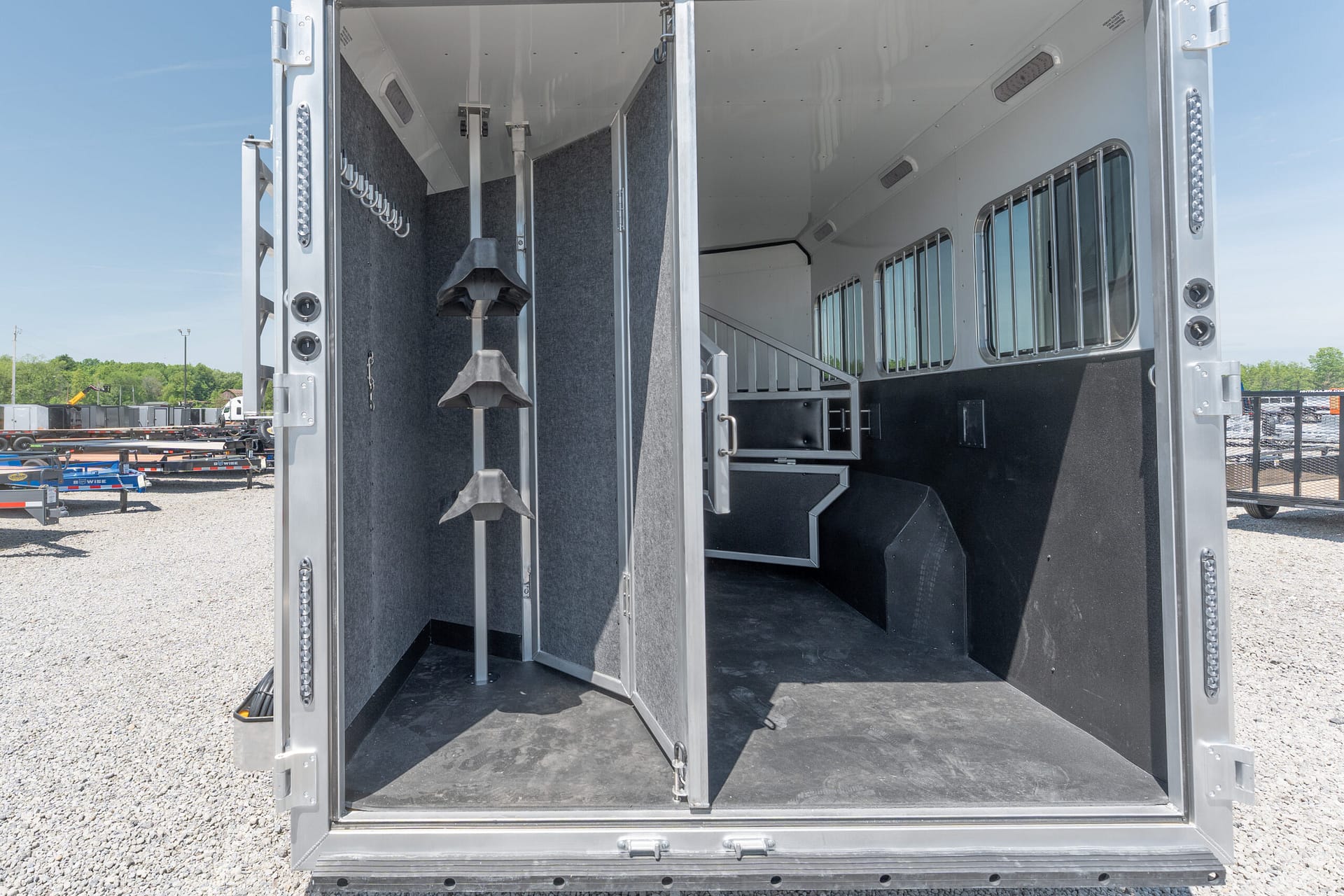 Open horse trailer interior with partitioned stalls, saddle racks, and bright daylight outside the open rear doors outside.