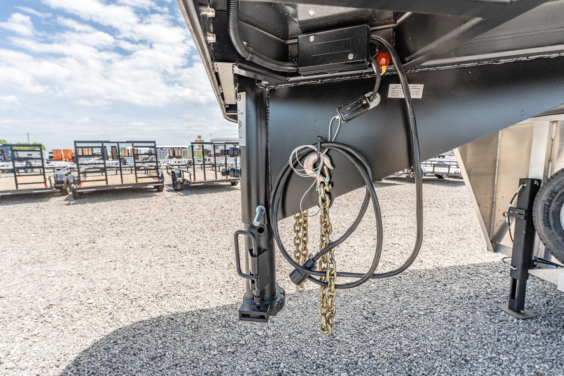 Close-up of a black trailer hitch with safety chains, electrical cables, and metal frame, on a gravel lot with other trailers in the background.] ,