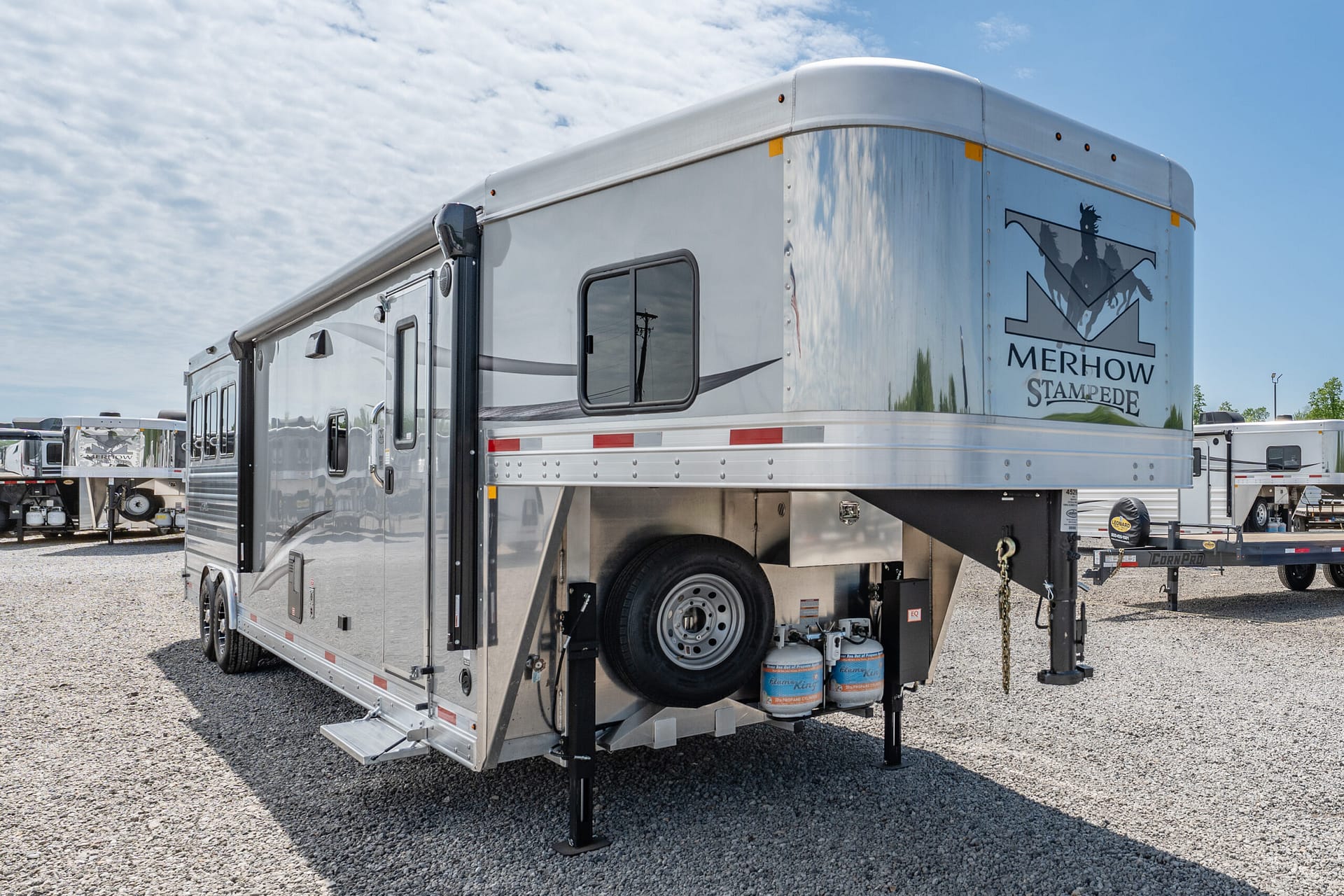 Silver horse trailer raised on its stabilizing legs in a gravel lot, Merhow Stampede logo on the rear panel.